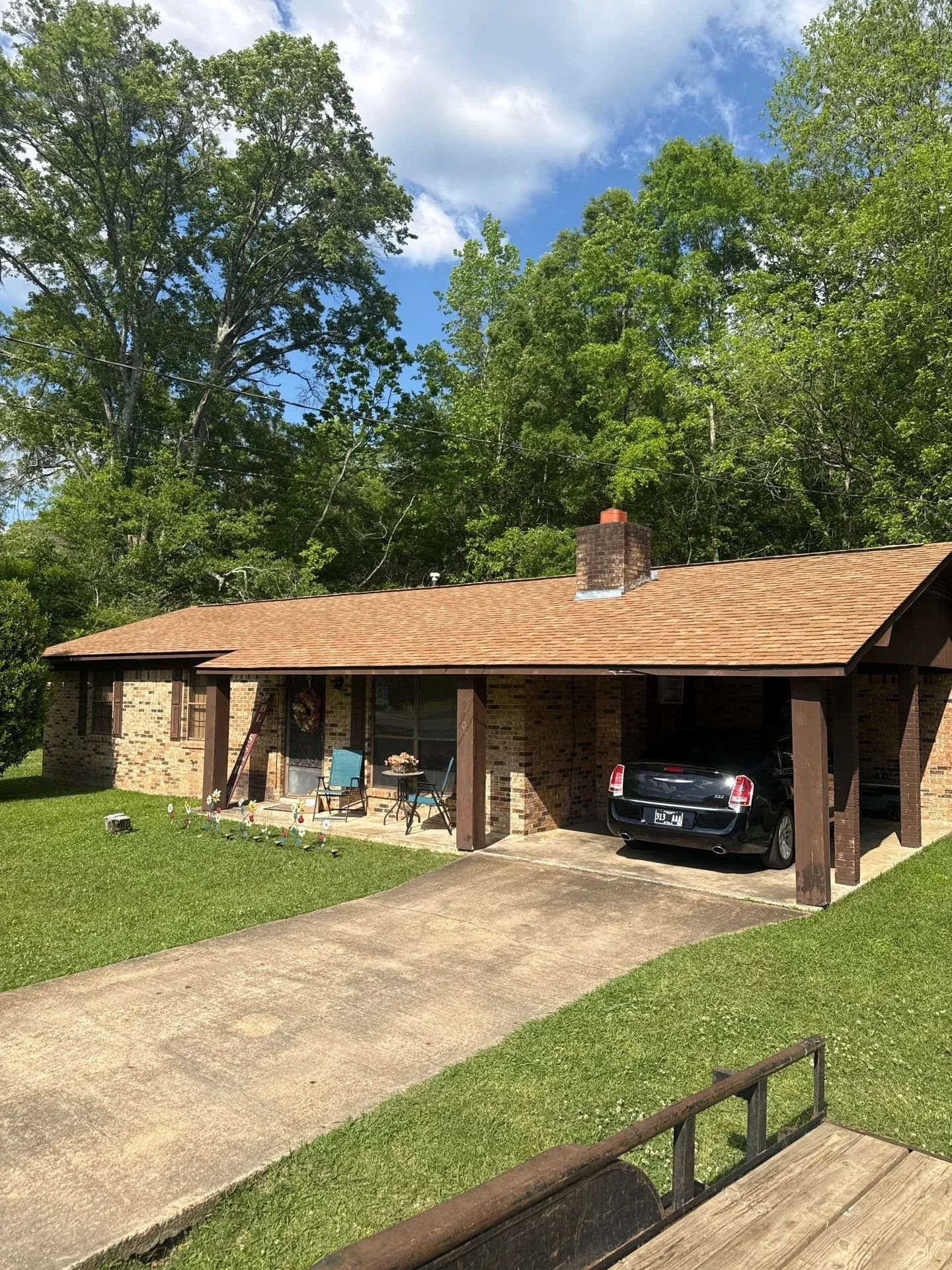 A brick house with a brown roof and a chimney, a driveway with a black car parked under a carport, a small patio with chairs and a table, a well-maintained lawn, and a forest background with green trees under a partly cloudy sky.