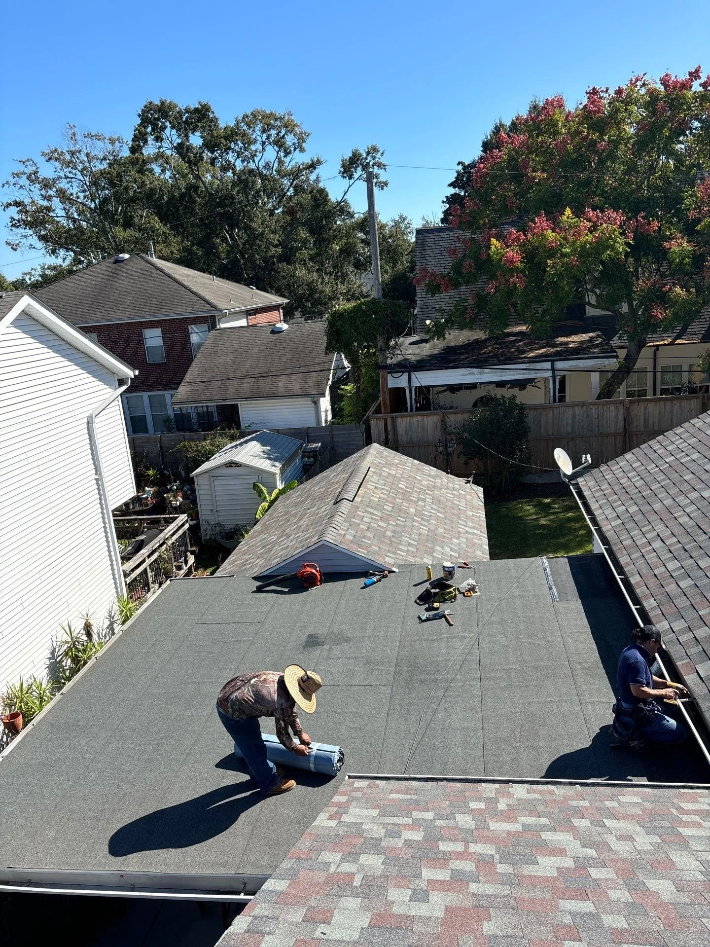 People installing a flat roofing system on a house, with tools and materials, on a sunny day with neighboring houses and trees in the background.