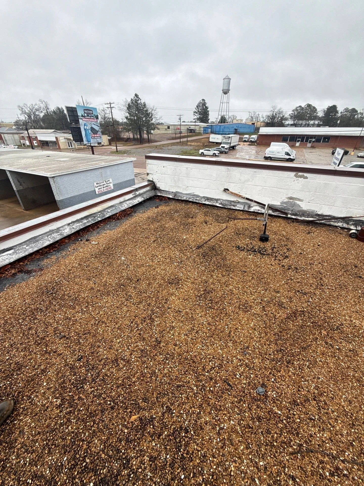 View from a rooftop showing a gravel surface, a white brick wall with damage, and a cityscape with vehicles and buildings in the background.