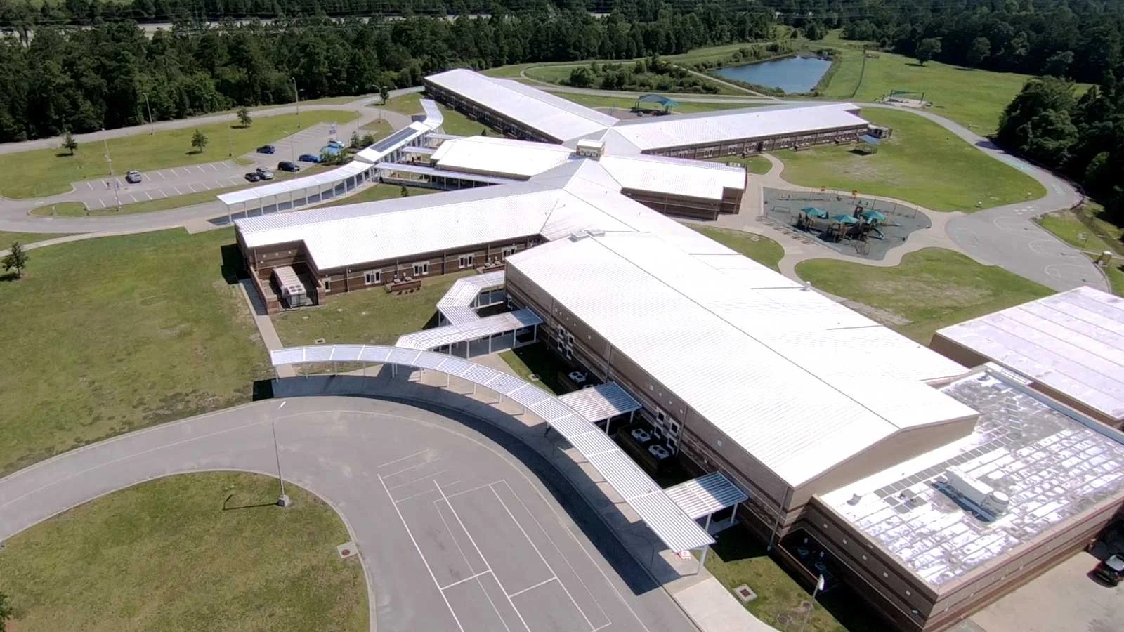 Aerial view of a school building with parking lot, playground, playground equipment, basketball courts, and a pond surrounded by trees.