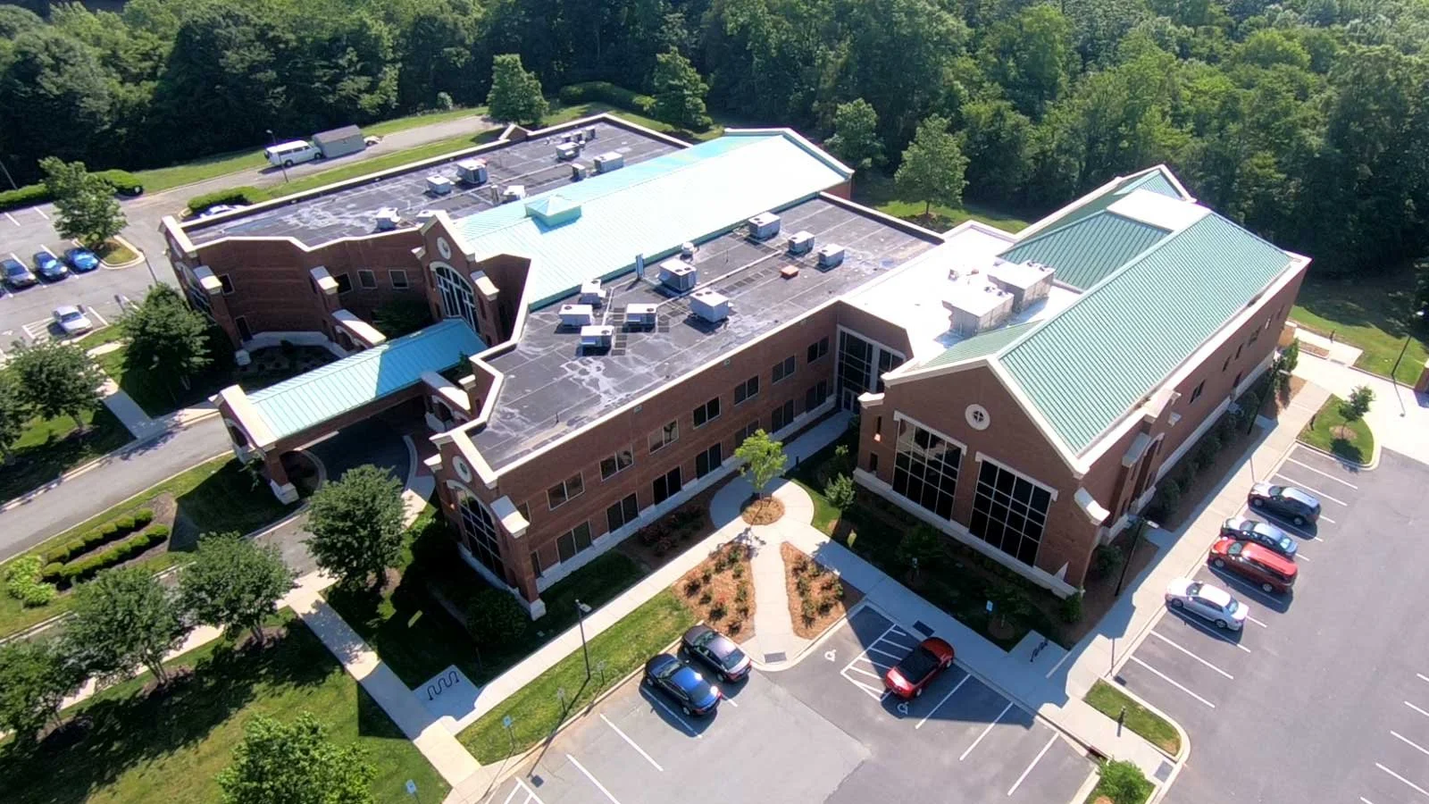 Aerial view of a large, modern building with a green metal roof, surrounded by parking lots and landscaped areas, adjacent to a wooded area.