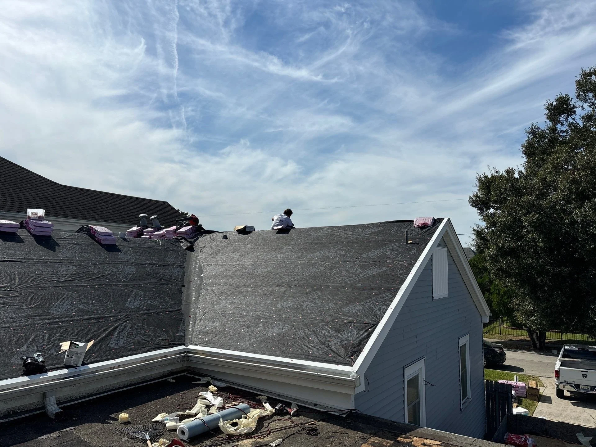 A house with a gray exterior and a steep roof under construction or renovation. The roof is partially covered with black underlayment, and a person is working on it. The sky is blue with scattered clouds, and there is a large tree to the right of the