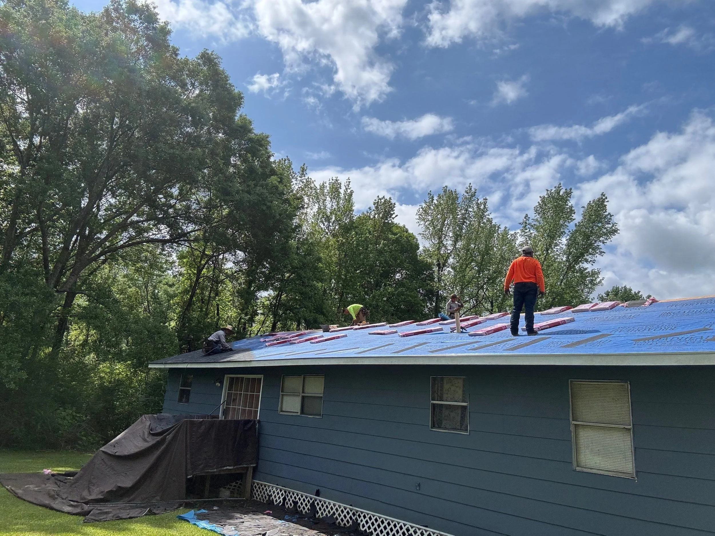 Workers installing new roofing on a blue house, with some tools and materials visible, surrounded by trees and a partly cloudy sky.