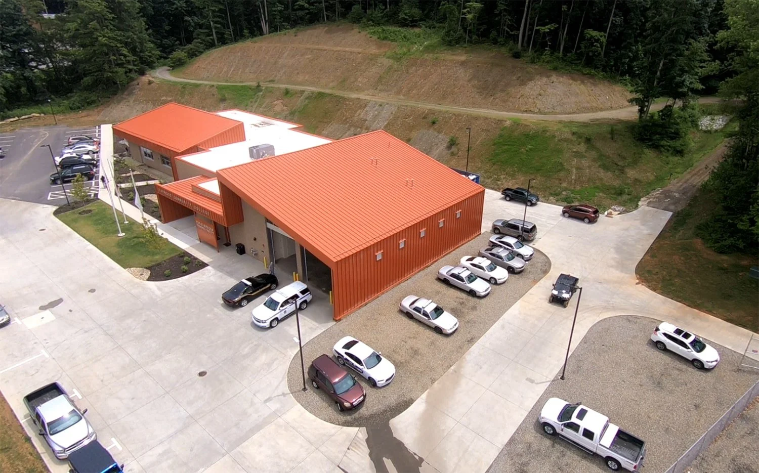 Aerial view of a modern building with orange metal roof and exterior walls, surrounded by a parking lot with cars, and a grassy hillside with trees in the background.