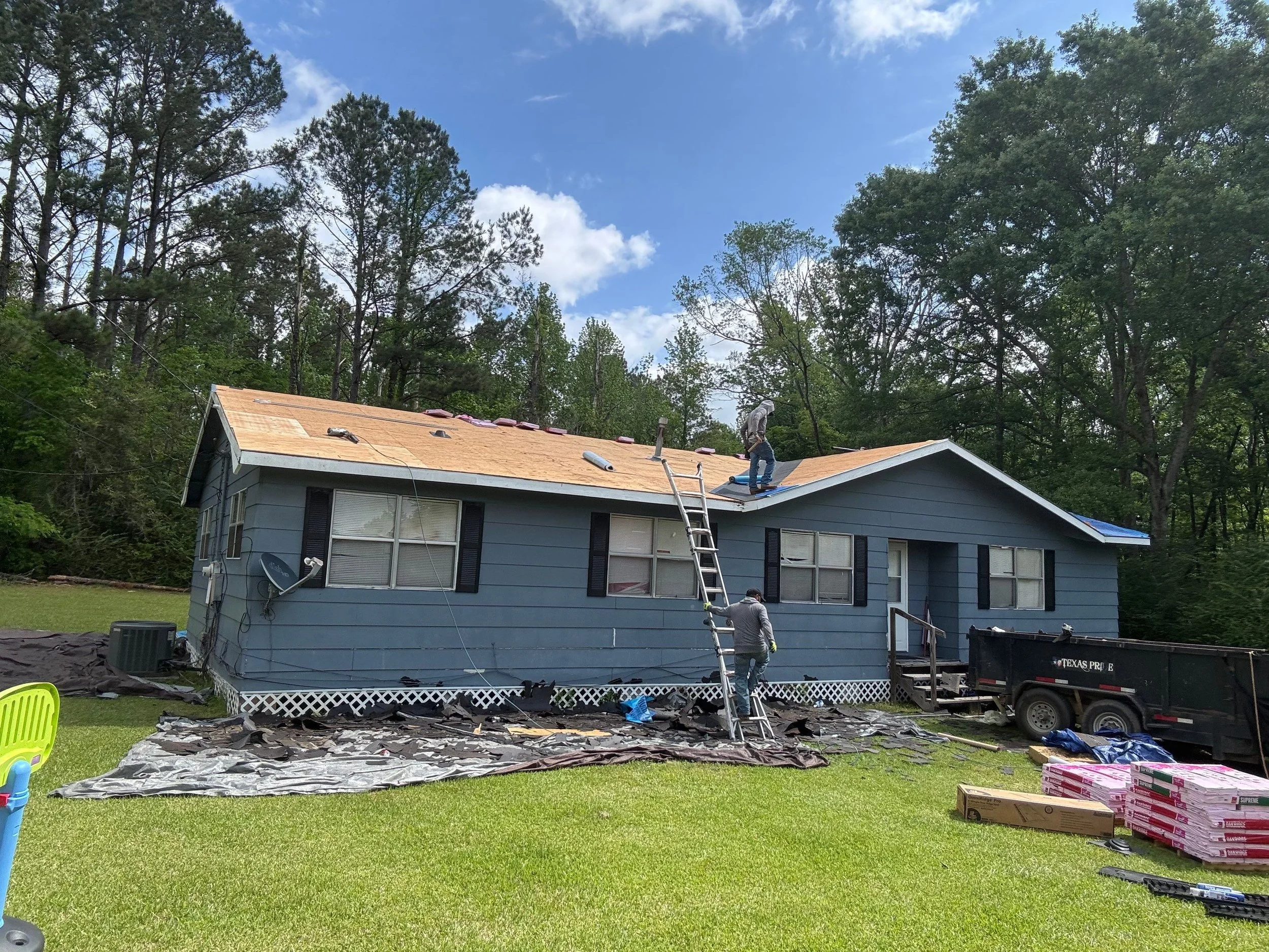 A house with its roof being renovated, with construction workers on the roof, ladder, construction debris and supplies on the ground, surrounded by trees and a partly cloudy sky.