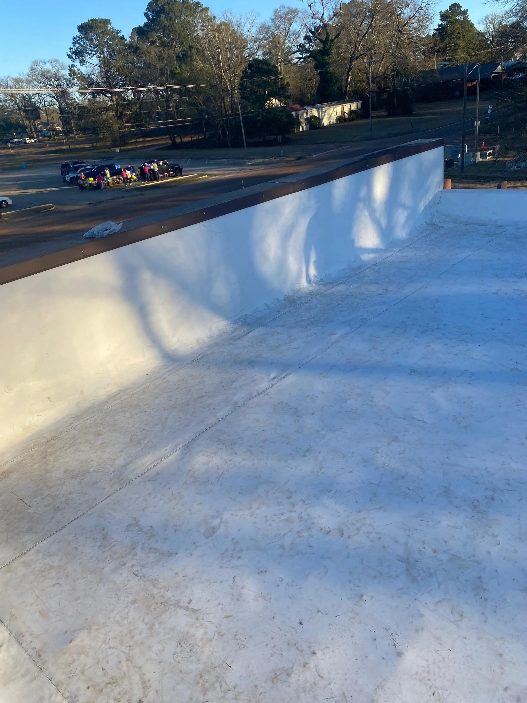 View from the top of a skateboarding ramp or ledge. There are shadows of people and trees cast on the surface of the ramp, with a parking lot and trees in the background.