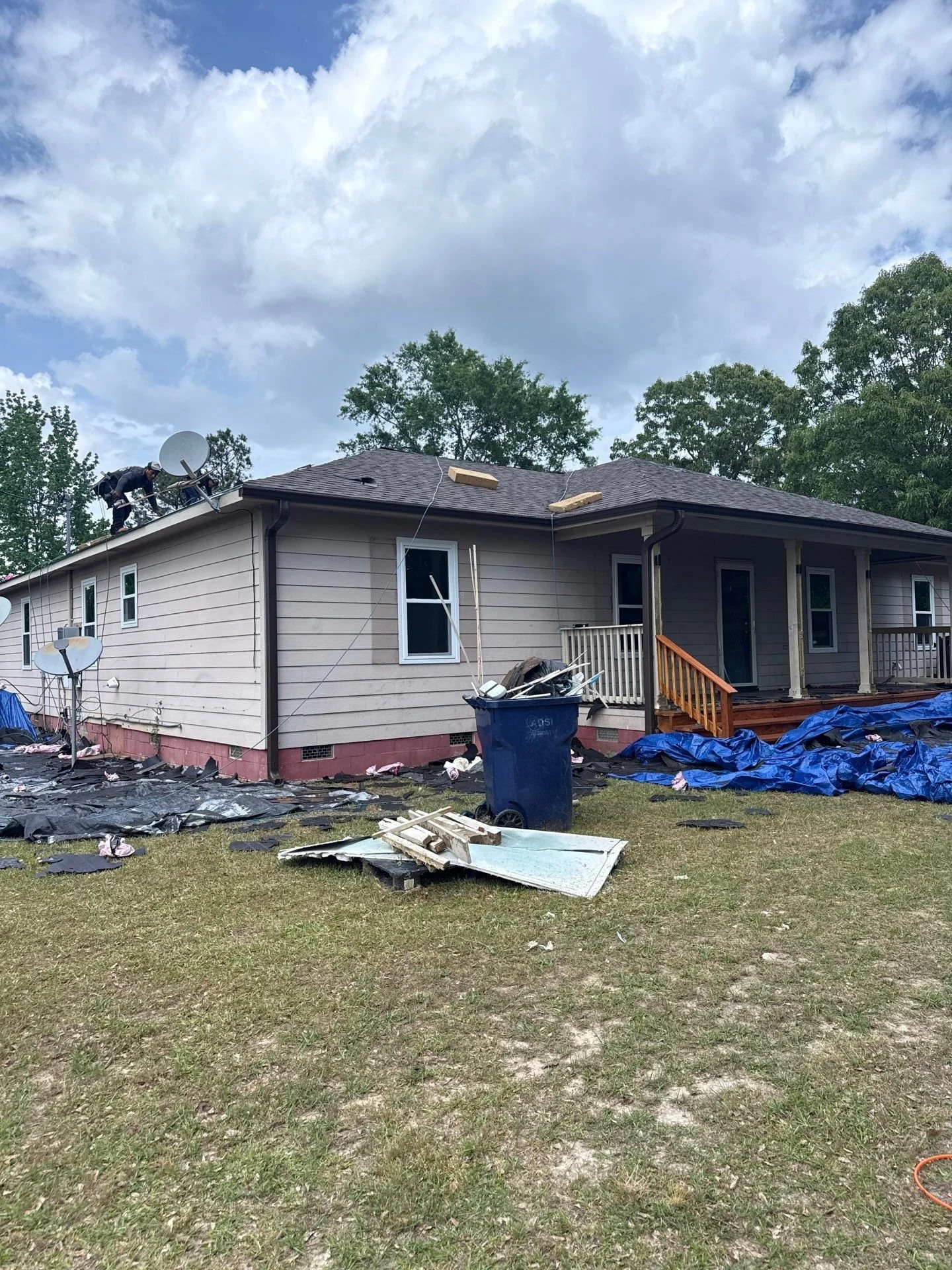 House undergoing roofing repairs with workers on the roof and debris scattered on the lawn