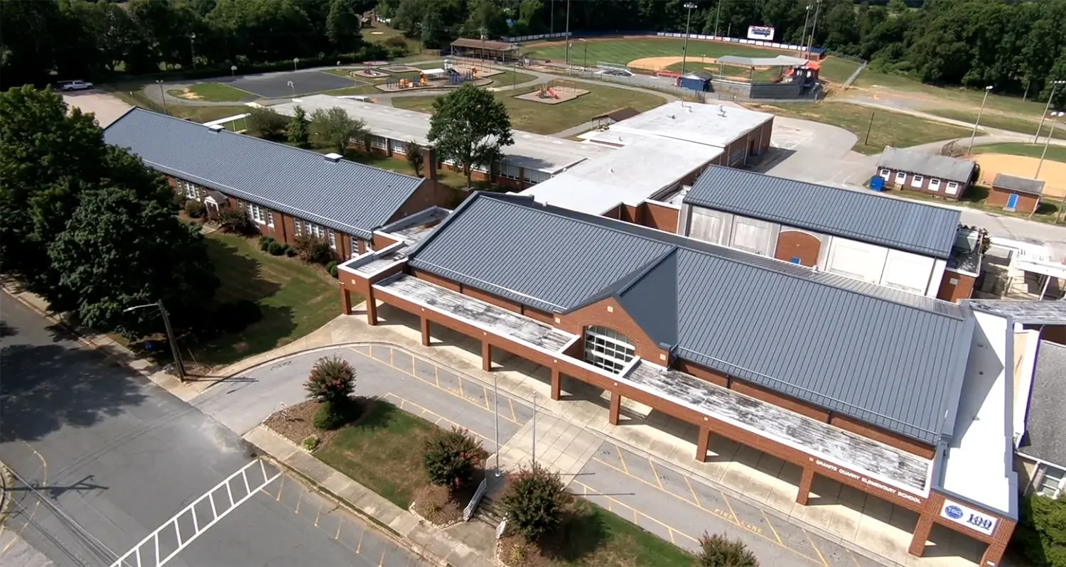 Aerial view of a school campus with multiple buildings, outdoor sports courts, playgrounds, and parking areas surrounded by trees.