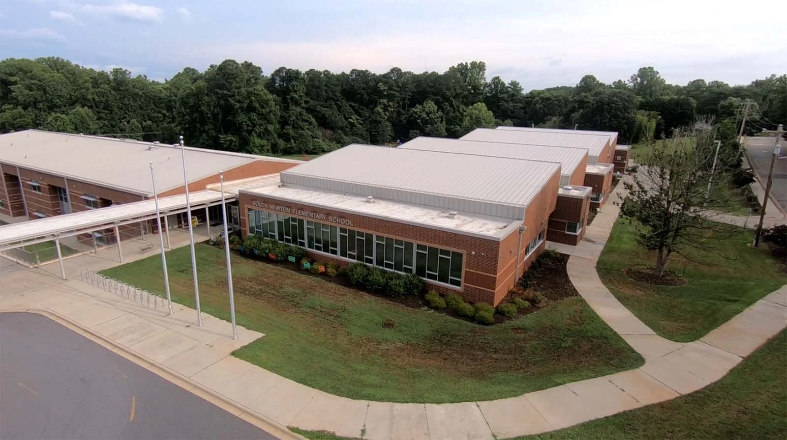 Aerial view of South Newton Elementary School, brick building, large windows, green lawns, trees, sidewalk, flagpoles, and nearby parking lot.