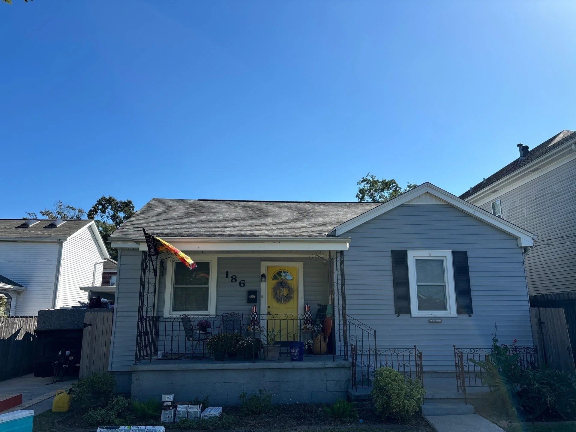 A white house with black shutters and a yellow front door, decorated with a holiday wreath. The house number is 186. The front porch has decorative black metal railings with potted plants and holiday decor. The house is in a neighborhood with similar