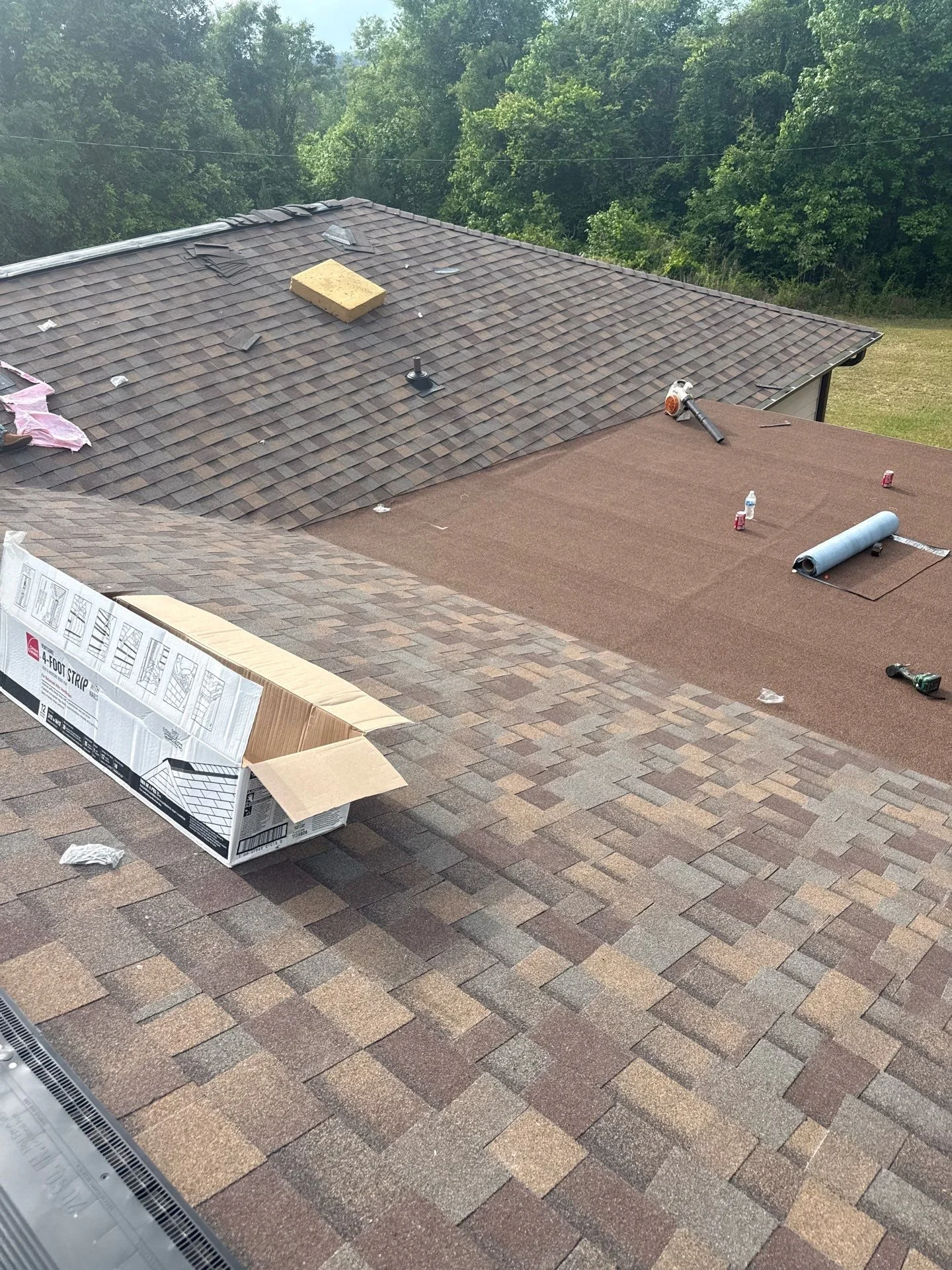 The image shows a house roof with shingles in the process of repair or replacement. There is an open cardboard box, a roll of roofing underlayment, a utility knife, a water bottle, and some red soda cans on the roof. The background features trees and