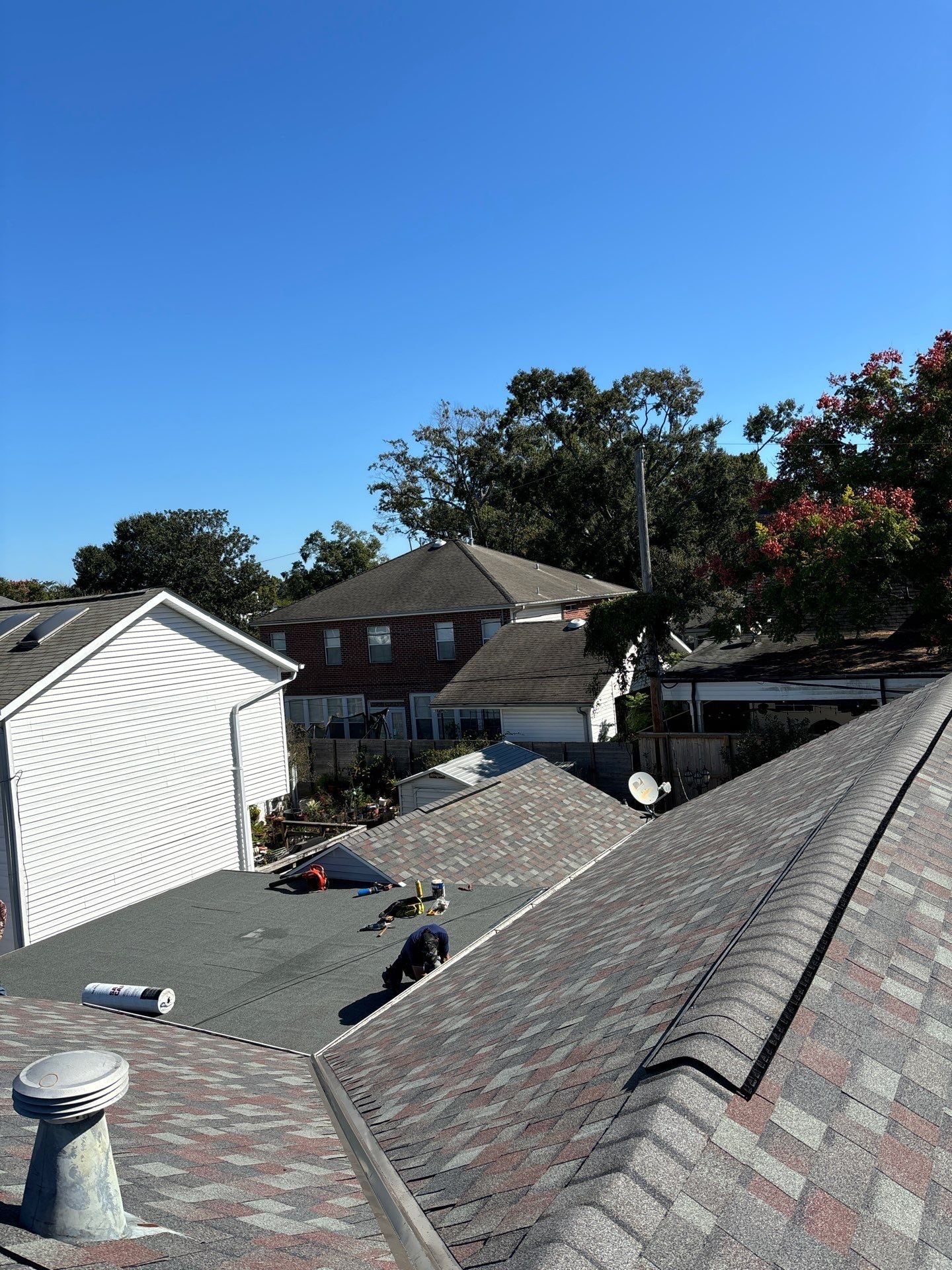 View of rooftops in a residential neighborhood with a clear blue sky, some houses with shingled and metal roofs, and workers on the roofs performing maintenance or repairs.