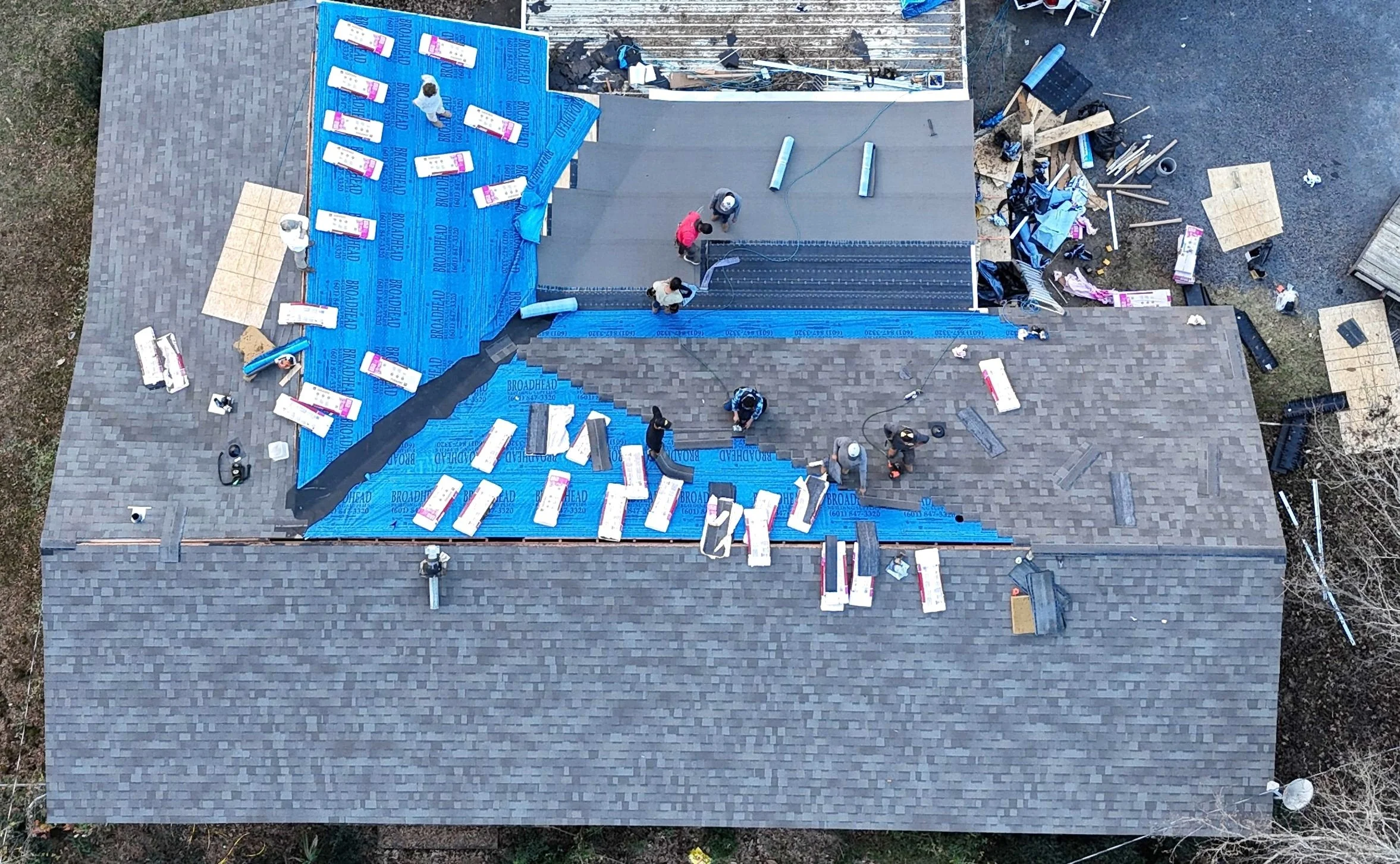Aerial view of a house roof under construction, with workers installing roofing materials, blue underlayment, and tools scattered around.
