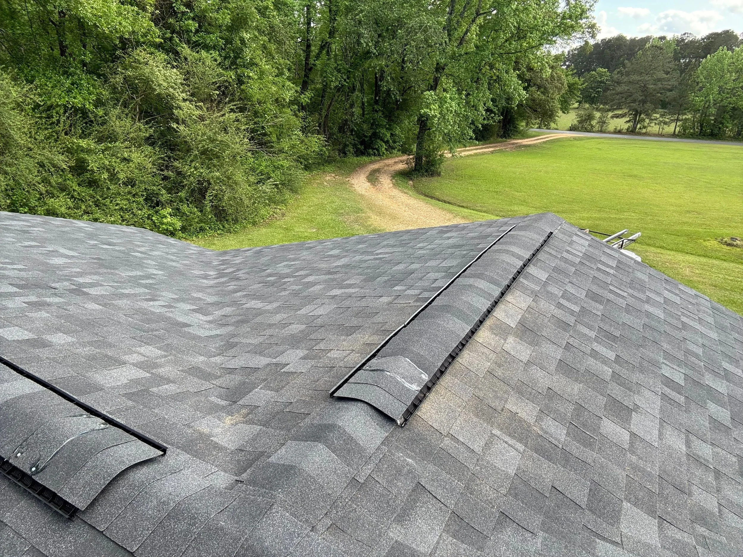 View from the roof of a house showing gray shingles, a green lawn, a dirt path, and a dense cluster of green trees under a partly cloudy sky.