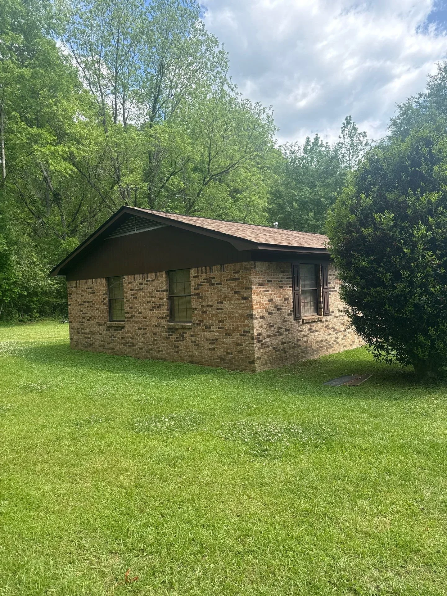 A brick house with four windows and brown shutters, sitting on a well-kept green lawn, with tall trees and a partly cloudy sky in the background.