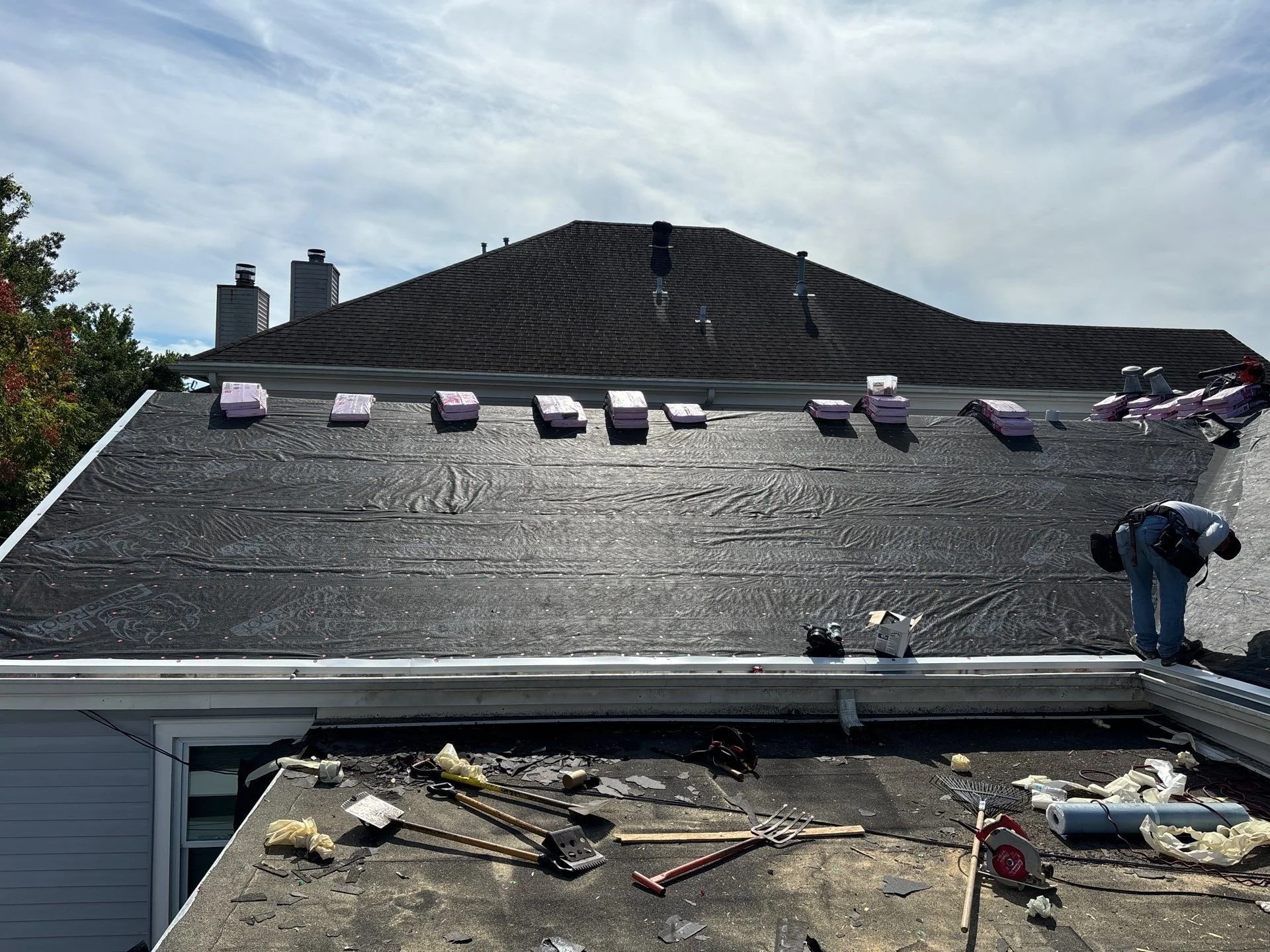 A house roof under maintenance with several tools, materials, and supplies scattered on the work area, and a worker in safety gear installing or repairing shingles amid daytime sky.