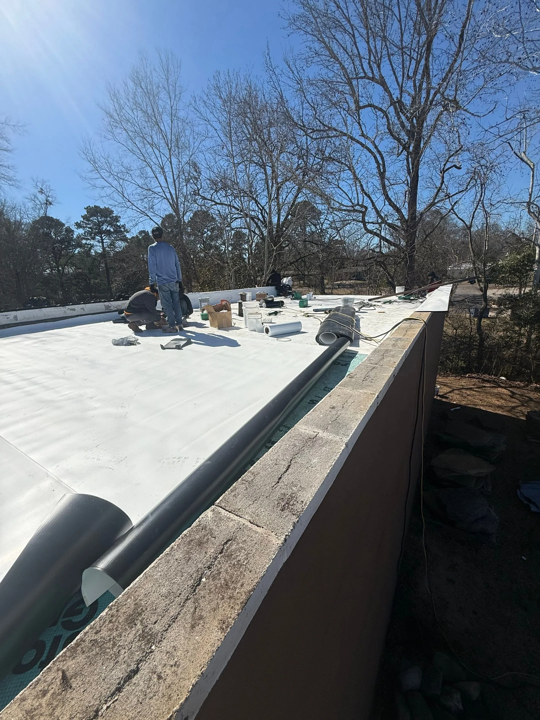 Construction workers are working on a flat roof, installing a weatherproofing material, with tools and materials scattered around.