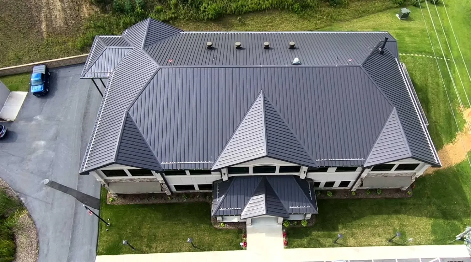 Aerial view of a large house with a dark metal roof, surrounded by green lawn and a parking area with cars.