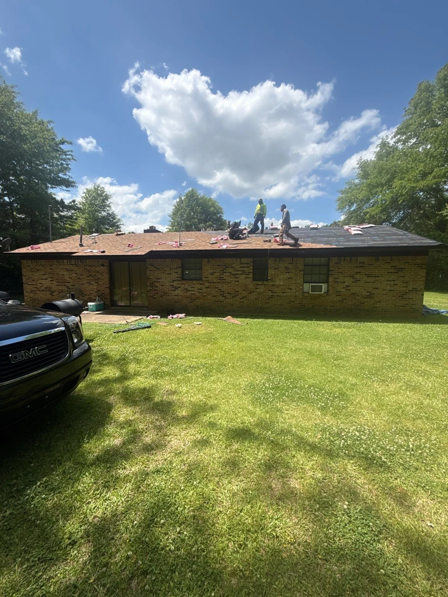 People working on the roof of a brick house on a sunny day with trees and a blue sky with clouds.