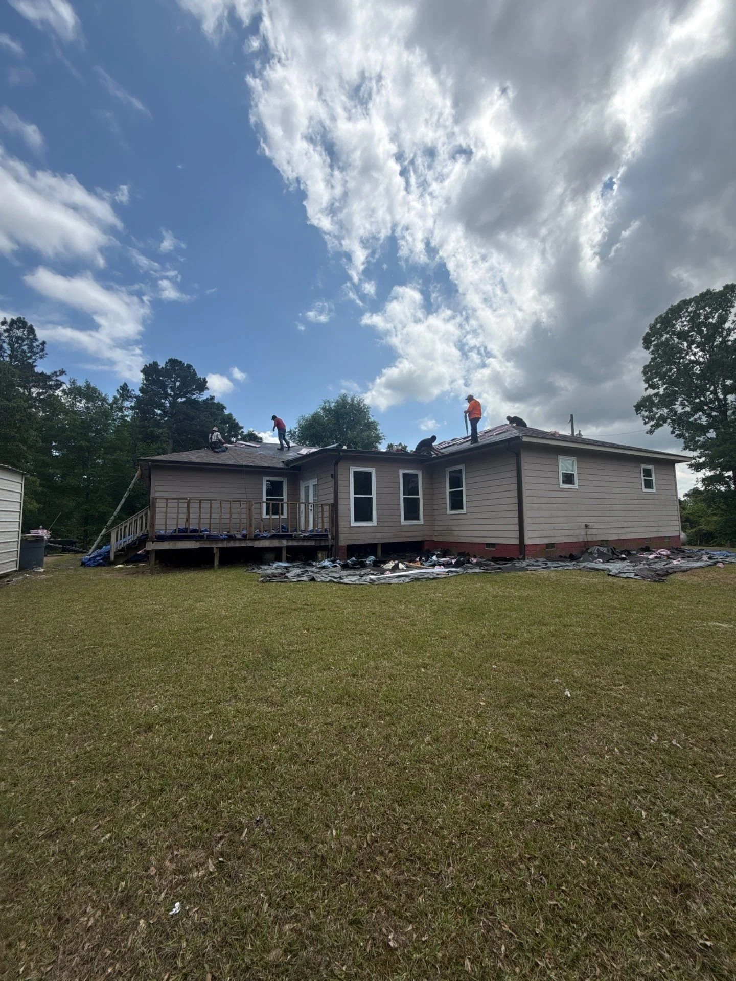 People working on the roof of a house during daylight, surrounded by tall trees and a partly cloudy sky.