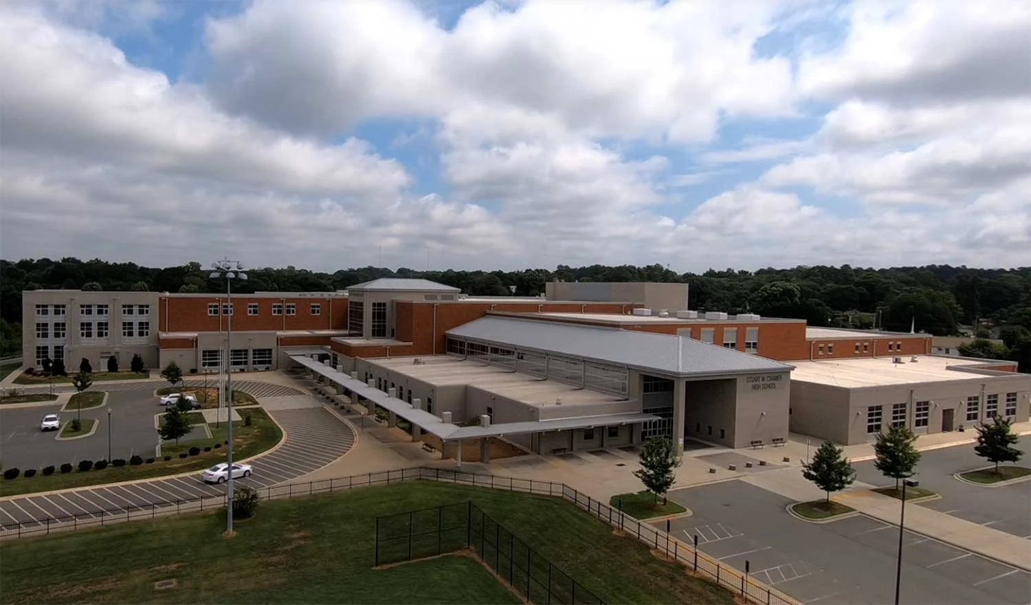 Aerial view of a high school building with parking lots, trees, and a green field, under cloudy skies.