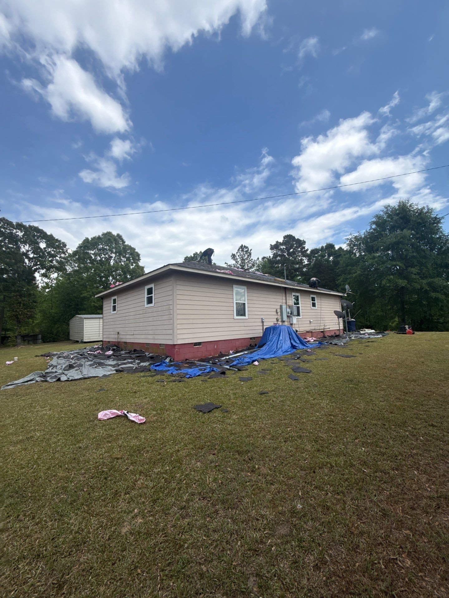 A house with damage to the roof, debris and tarps on the ground, and a partly cloudy sky above.