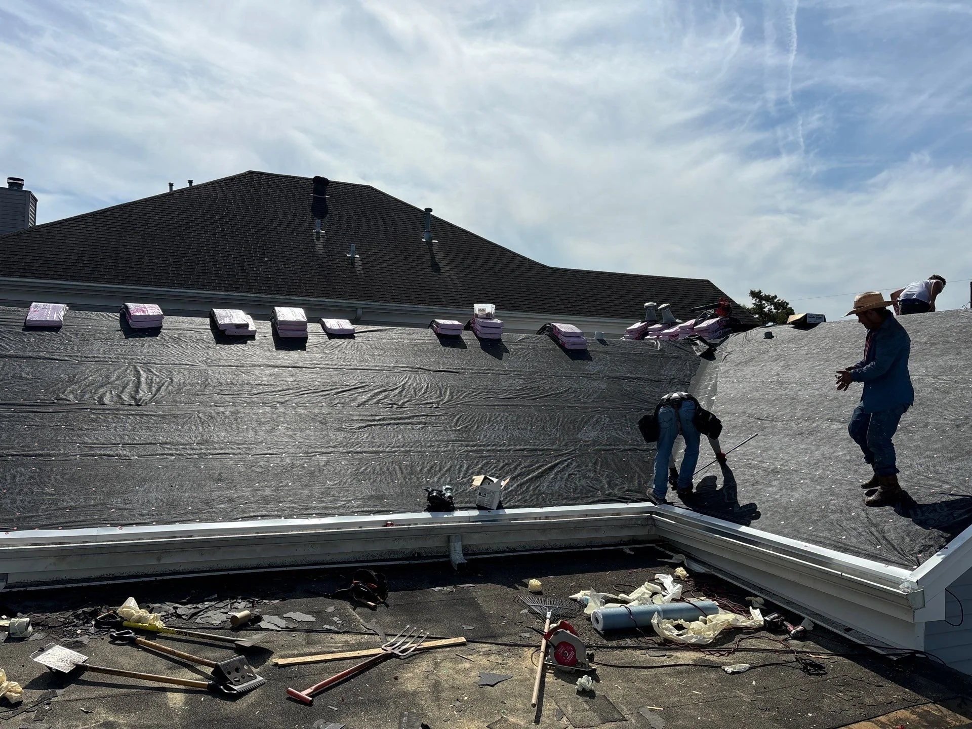 Roof repair workers replace shingles on a house roof, with tools and materials scattered around, under a cloudy sky.