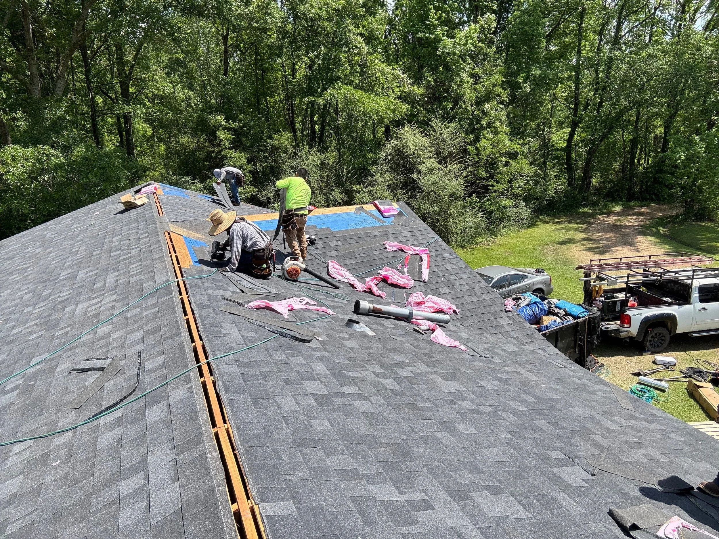 Three workers installing new shingles on a roof surrounded by green trees, with construction tools and materials laid out on the roof.