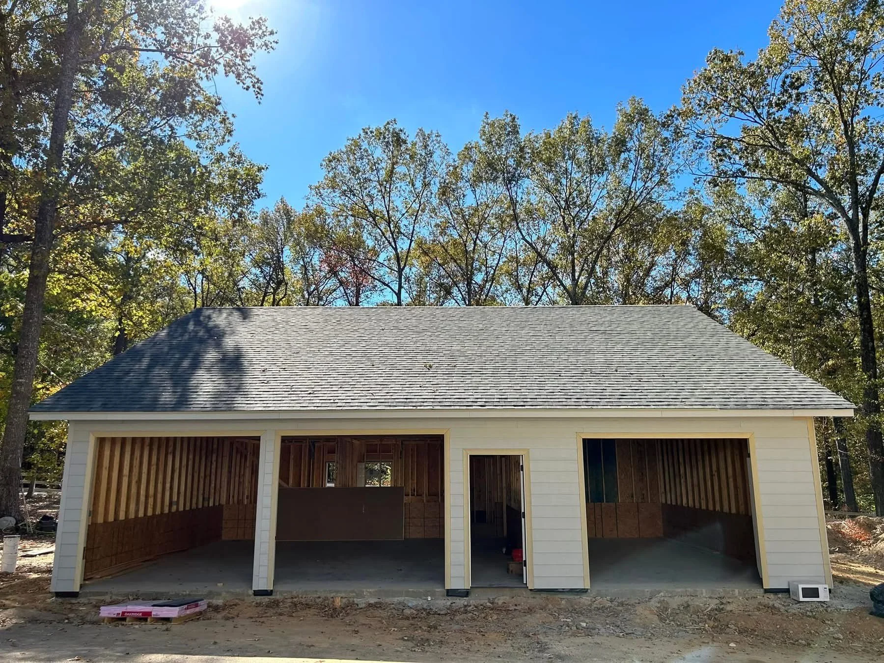 under construction garage with an unfinished interior, white siding exterior, and a gray shingle roof, set against a backdrop of trees and a blue sky