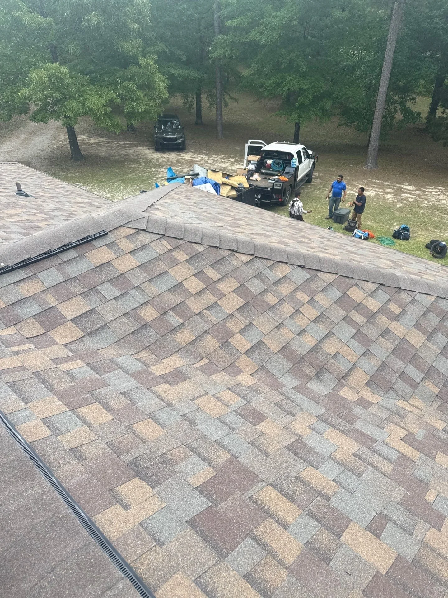 View from a high window showing rooftops with shingle shingles. In the background, a grassy yard with trees, a pickup truck, and a group of people standing near the truck and on the grass, surrounded by outdoor equipment and supplies.