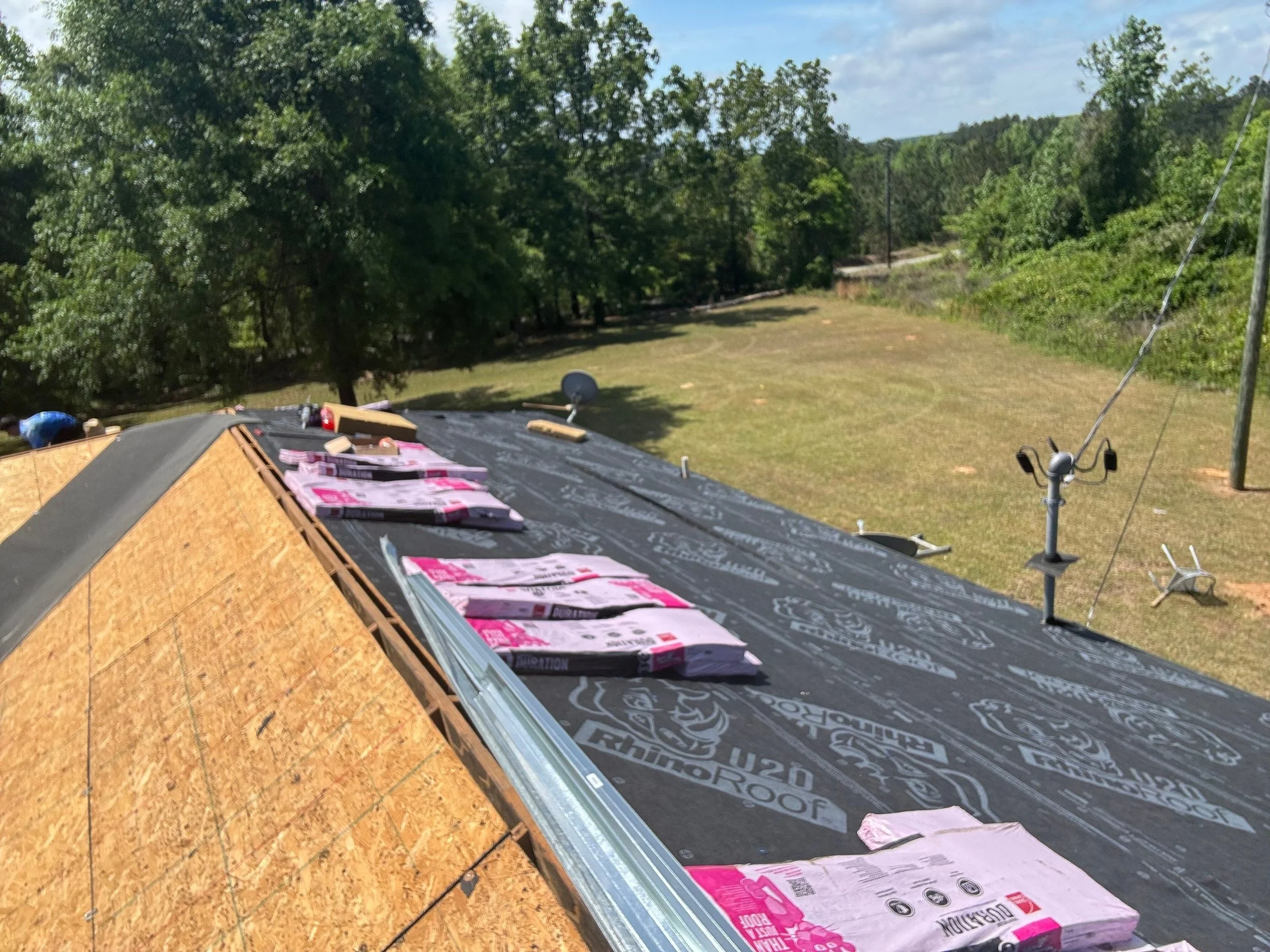 Construction site roof with black underlayment, pink insulation boards, and a partially installed wooden roof panel. In the background, there are green trees, a grassy area, and nearby power lines.