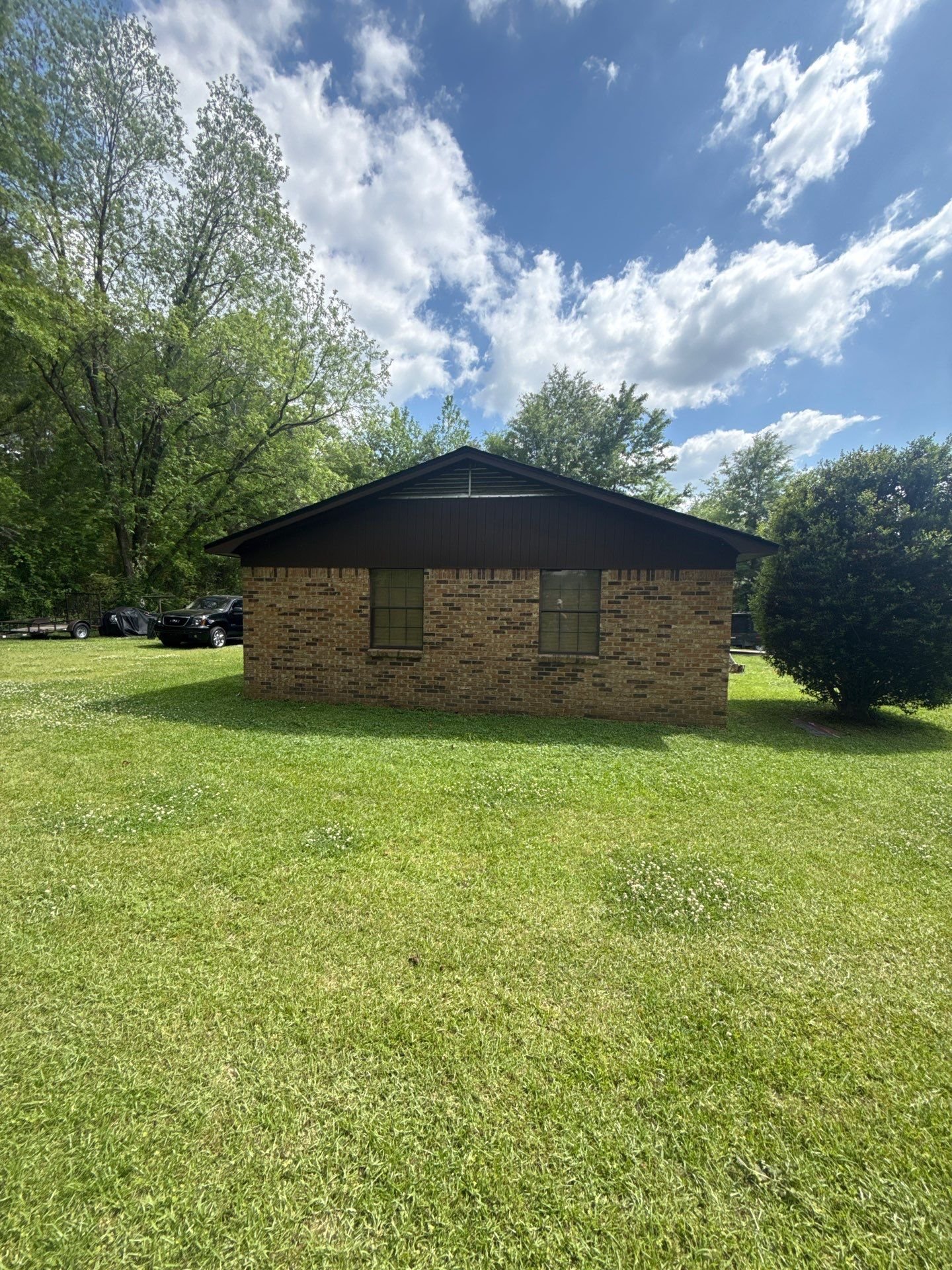 A small brick house with a black roof and two windows, surrounded by green grass and trees against a blue sky with white clouds.