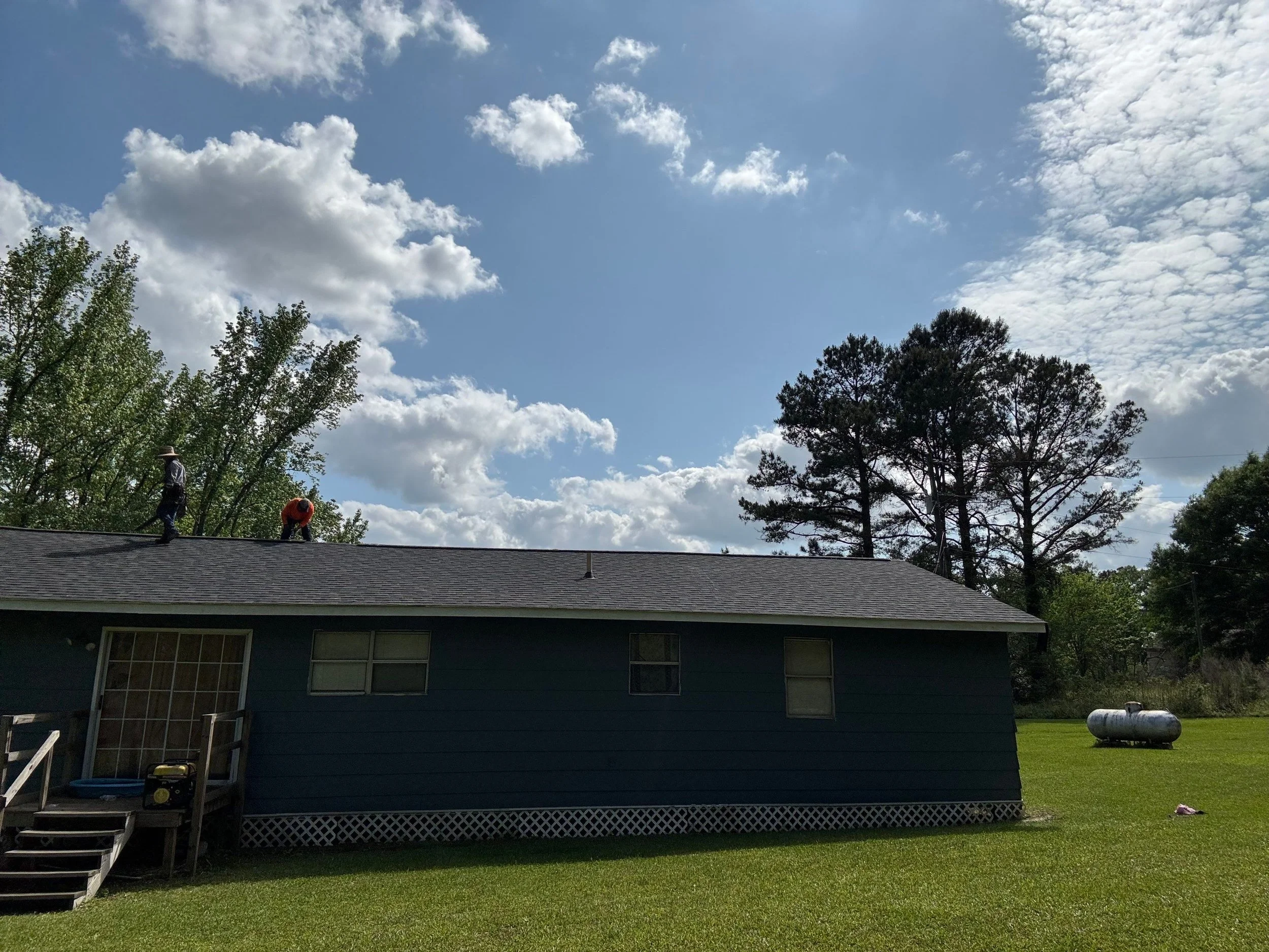 A blue house with a gray roof, front steps, and a large window, surrounded by green grass and trees. Two workers are on the roof, one bending down and the other standing. The sky is partly cloudy with white clouds and blue sky.