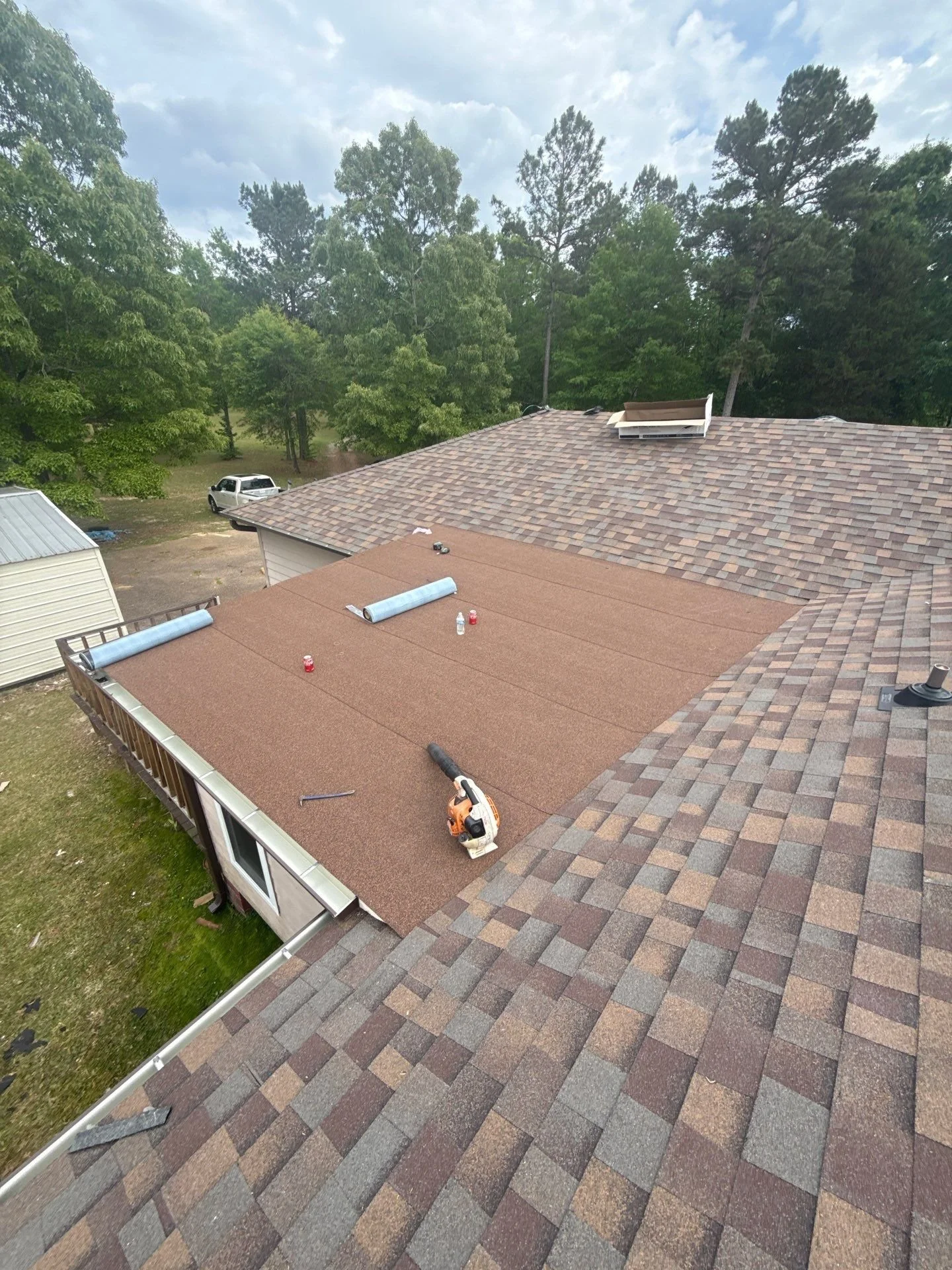 A house roof with new shingles being installed, with roofing tools and materials on the roof, surrounded by trees and a cloudy sky.