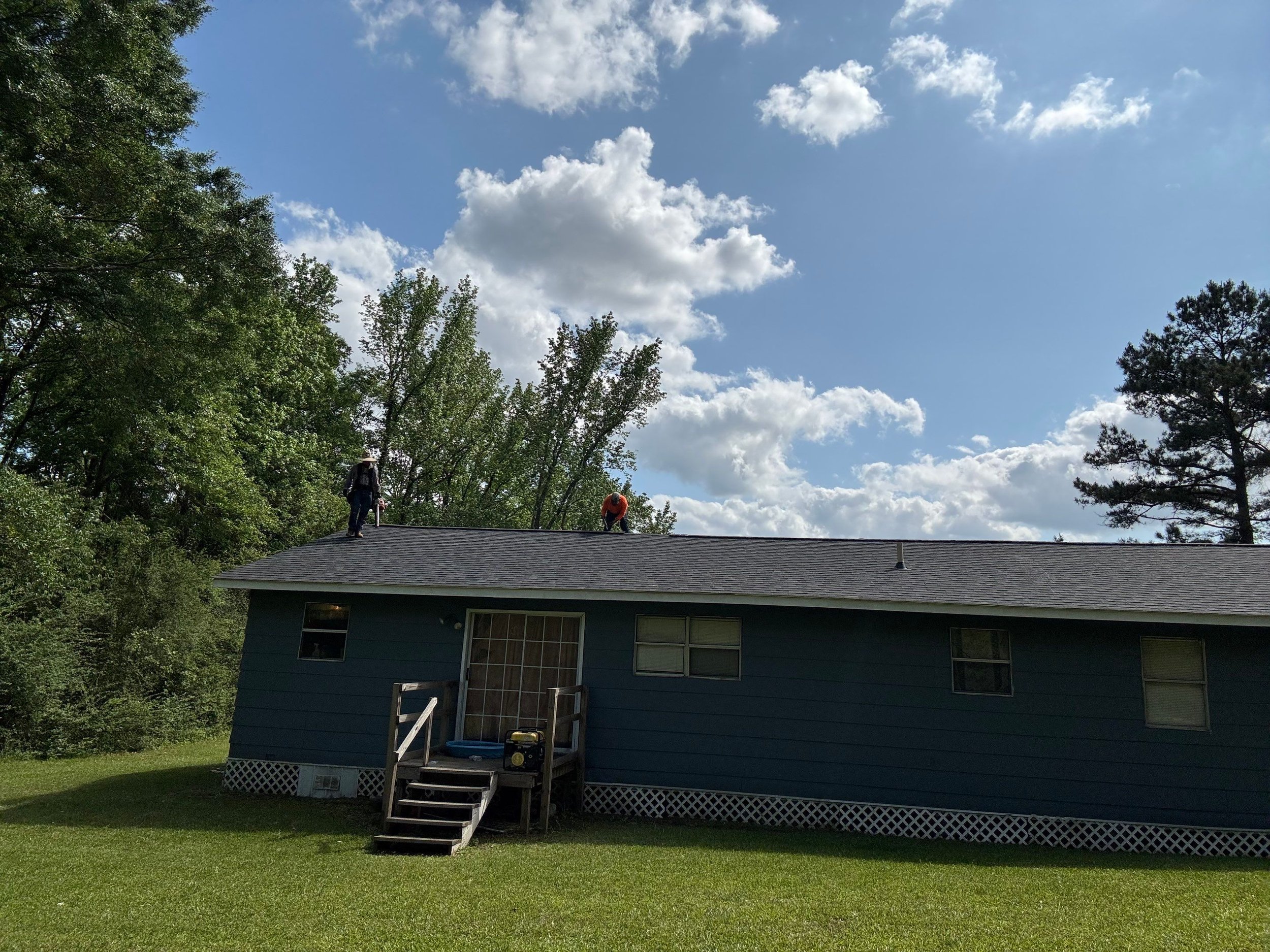 Two workers standing on a roof, repairing or inspecting it, with a blue house beneath and trees with green leaves around, under a partly cloudy sky.