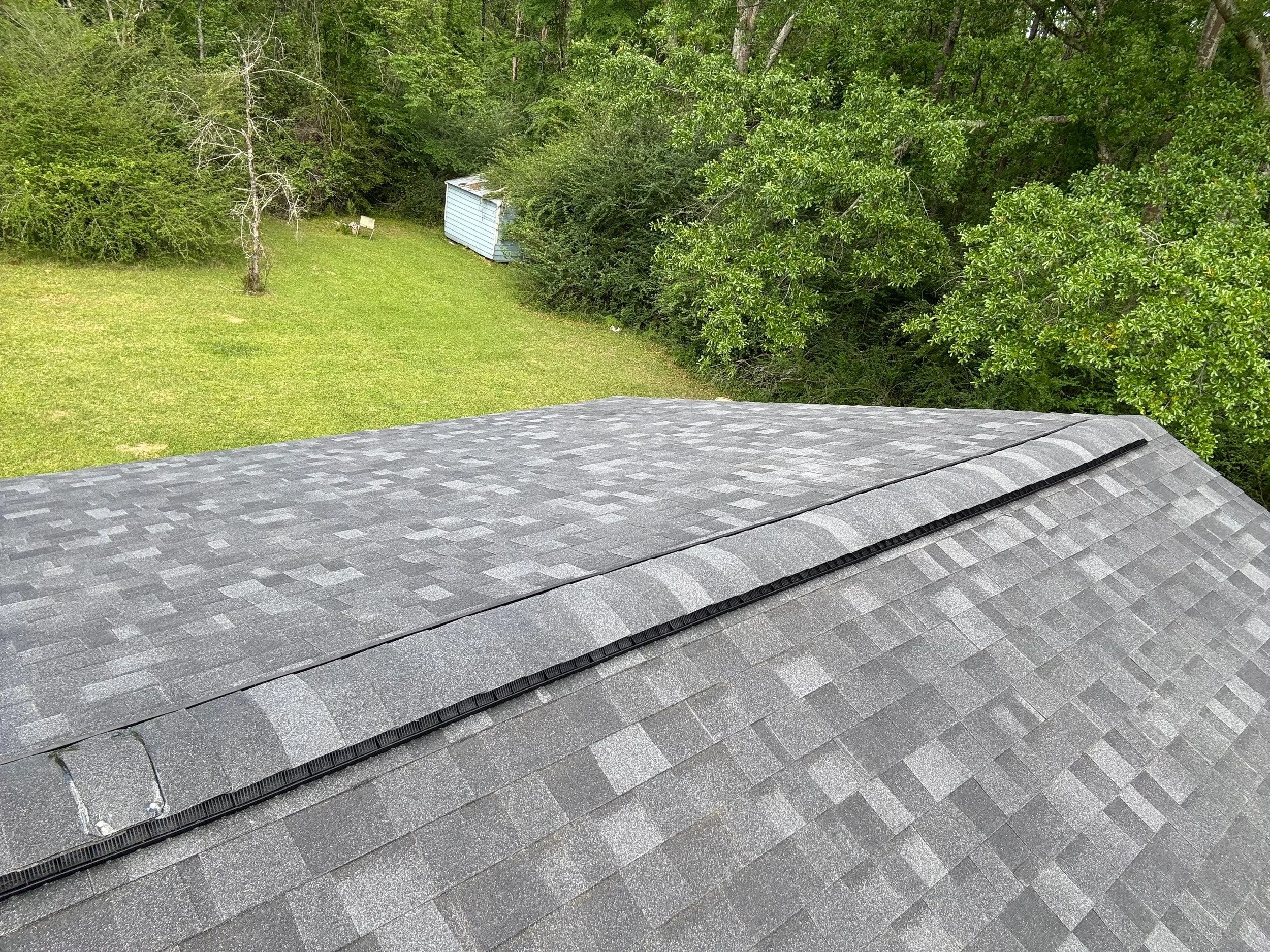 View of a rooftop with gray shingles, green trees, and a backyard with a green lawn, white chairs, and a small shed, seen from above.