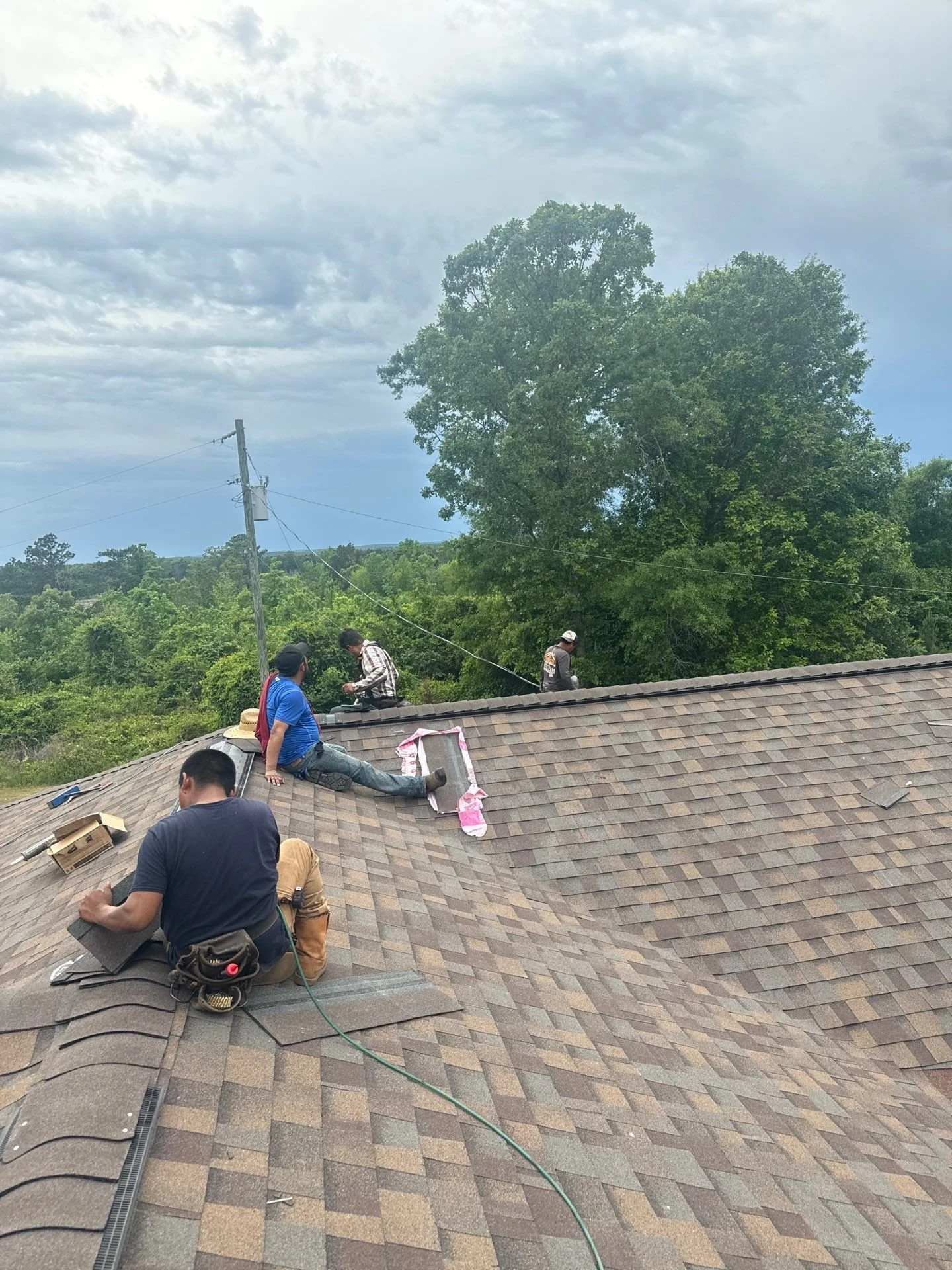 Roofers installing shingles on a residential roof during daytime, with trees and a cloudy sky in the background.