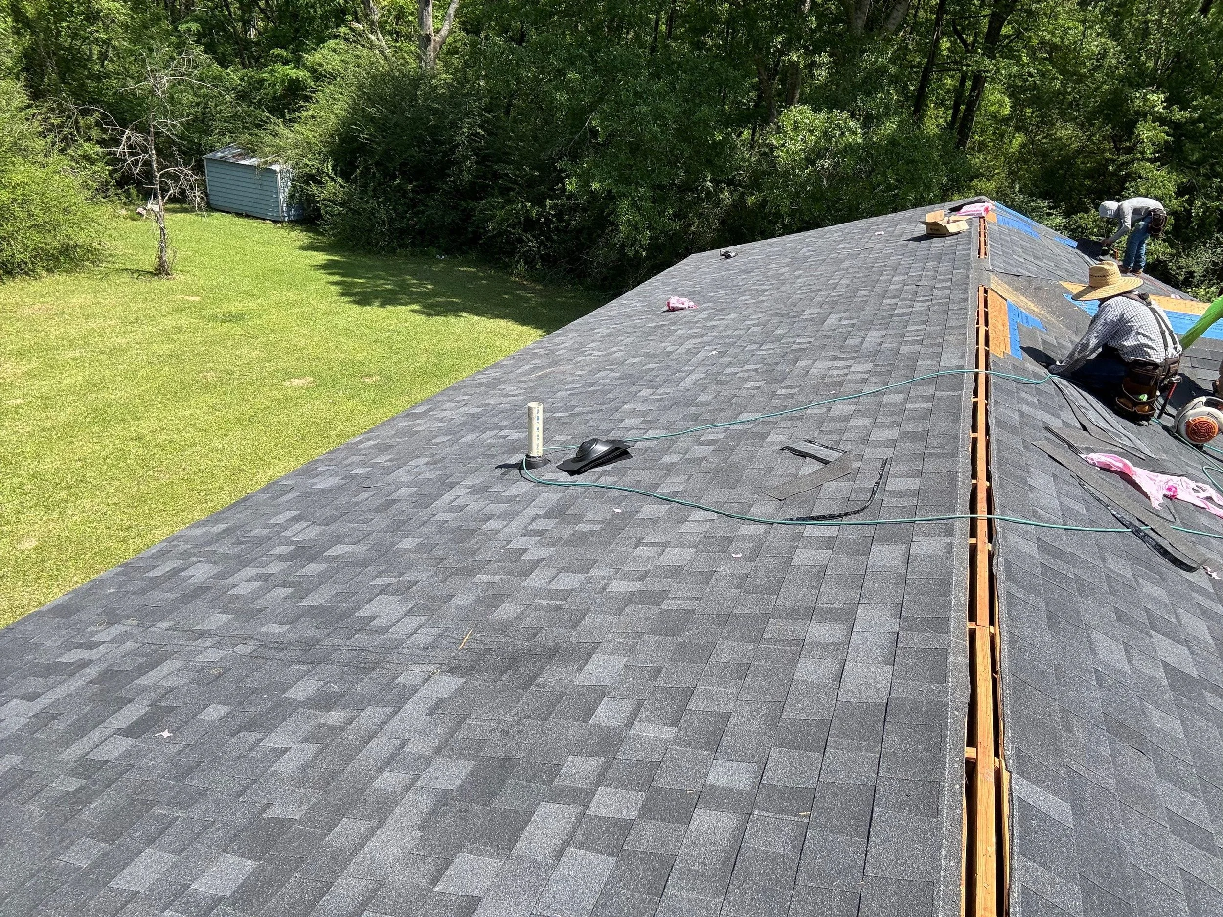 Roofing workers installing shingles on a house roof, with tools and materials nearby, surrounded by trees and a green lawn.
