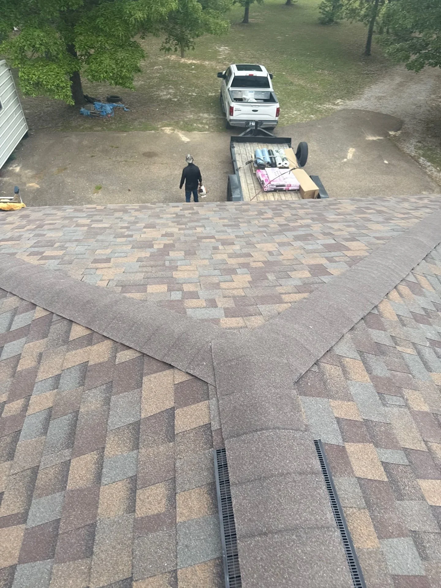 View from a rooftop looking down at a person walking on a driveway. There is a trailer with construction materials and a pickup truck at the end of the driveway, surrounded by trees and grassy areas.