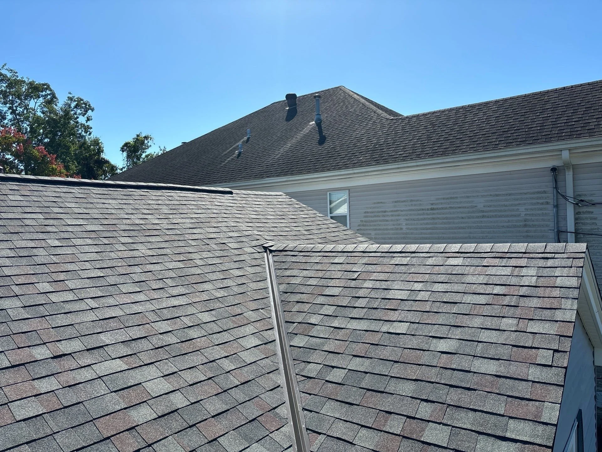 View of rooftops with asphalt shingles, a house wall with siding, a window, and some vents on the roofs under a clear blue sky.