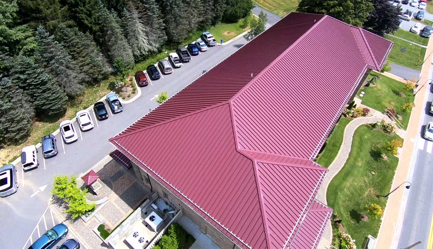 An aerial view of a building with a red metal roof surrounded by a parking lot with several cars, green lawns, trees, and brick pathways