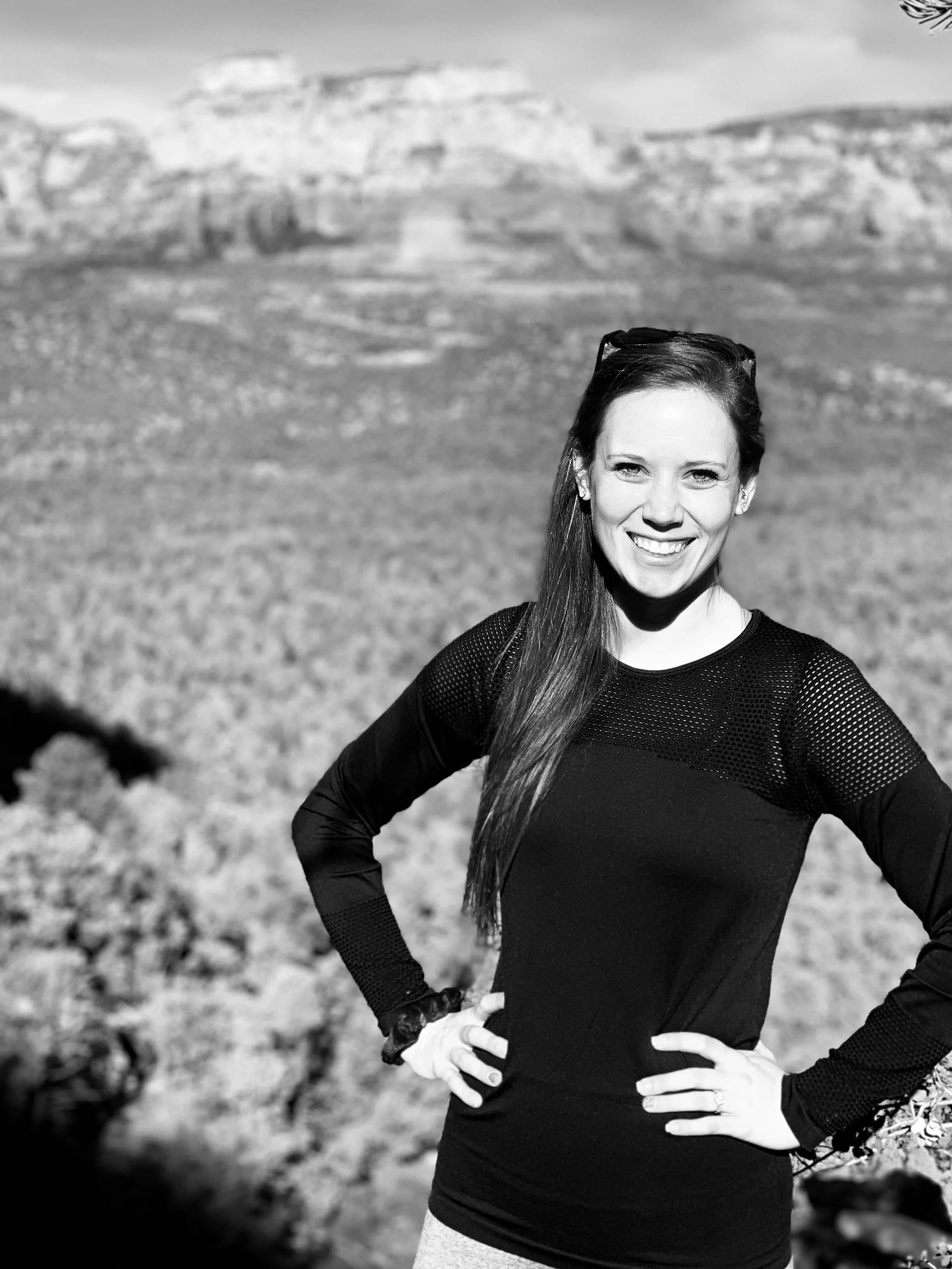 Black and white photo of a smiling woman standing outdoors with her hands on her hips, rocky terrain, and distant mountains in the background.