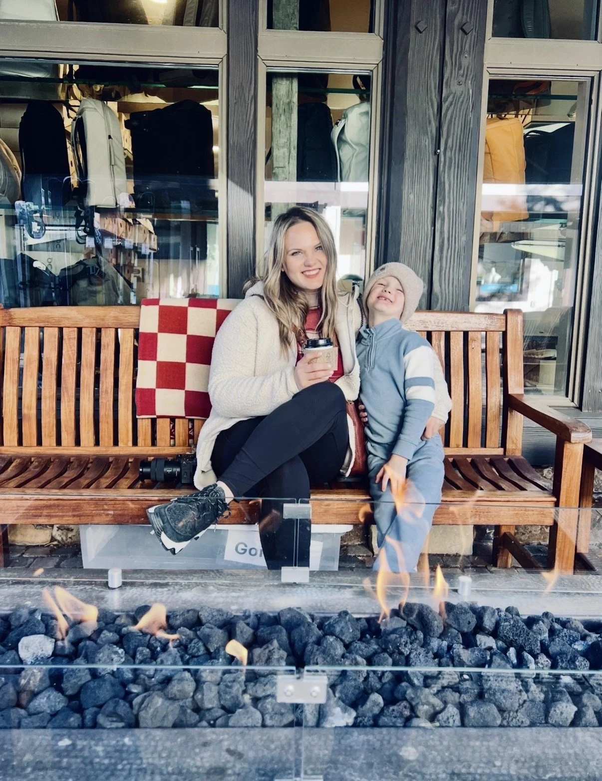 A woman and a young boy sitting on a wooden bench outside near a fire pit. The woman is smiling, holding a cup, and wearing a cream fleece jacket and black pants. The boy is leaning into her, smiling, wearing a gray beanie and a blue and gray hoodie with matching pants. Behind them is a large window with backpacks and bags visible inside.