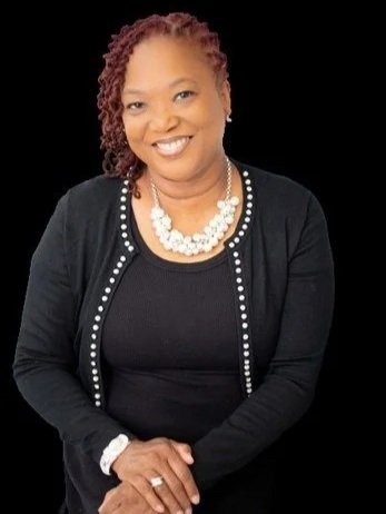 A woman with shoulder-length curly hair, wearing a black top with a black cardigan that has pearl accents, and a pearl necklace, smiling against a black background.