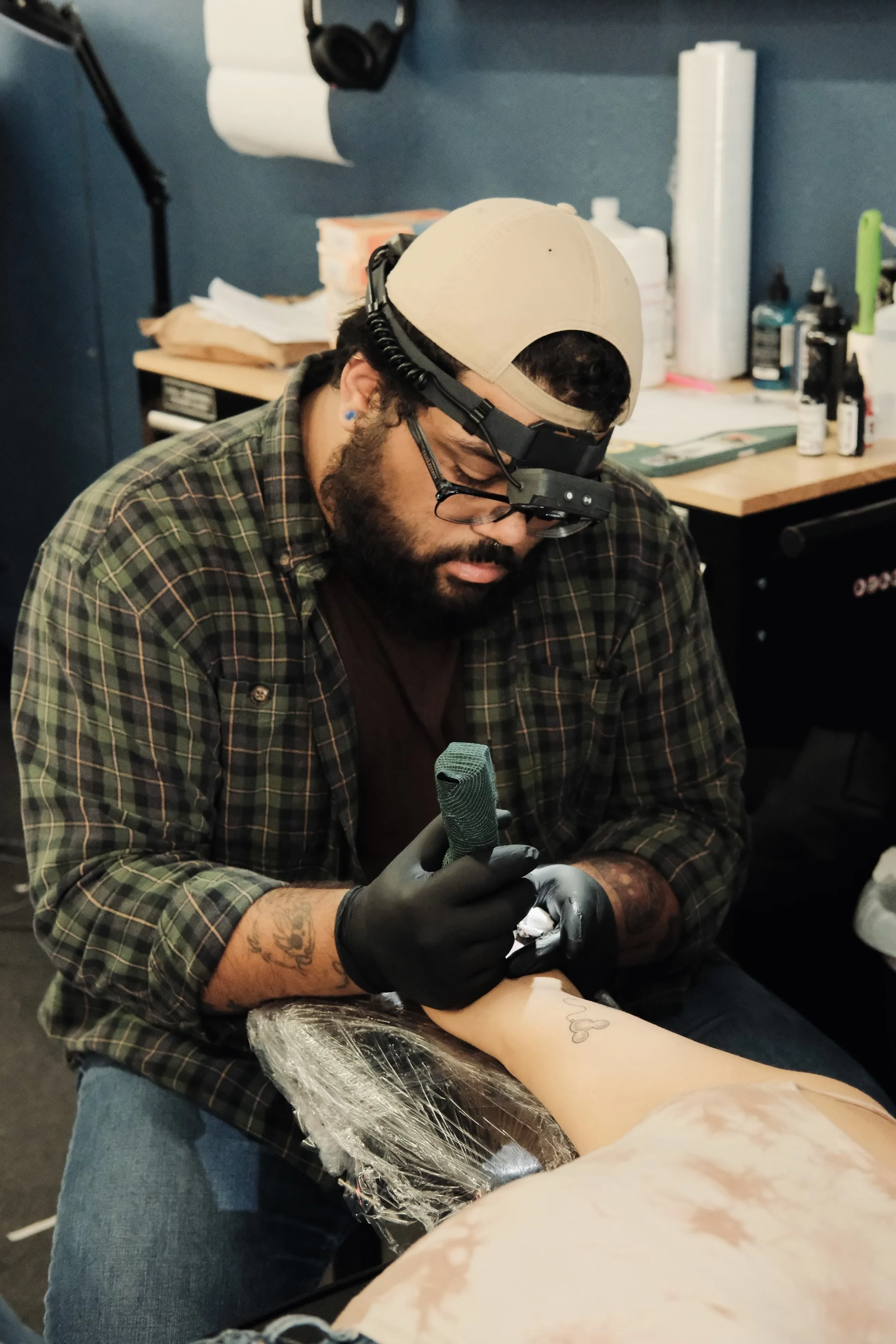 A man wearing glasses, a backward cap, and black gloves tattooing a person's arm with a tattoo machine in a tattoo studio.