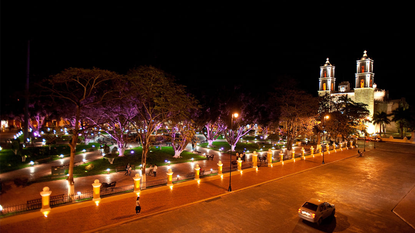 Night view of a park with trees illuminated by purple and pink lights, benches, walkways, and small decorative lights. In the background, there is a historic building with two towers lit up at night.