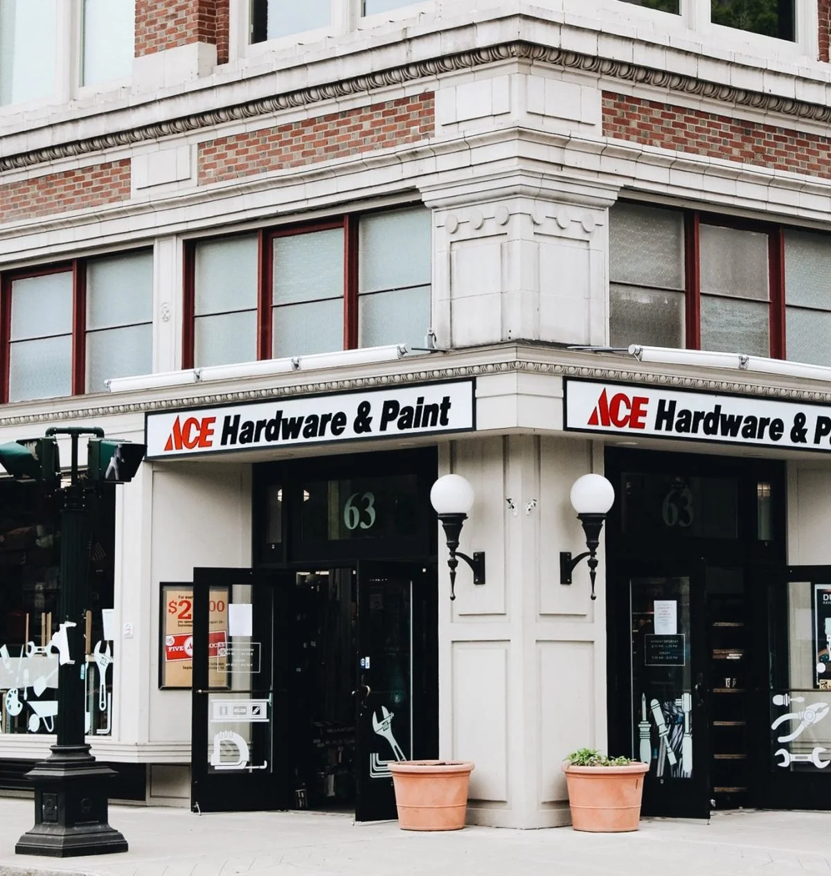 Corner of a storefront for ACE Hardware & Paint on a city street, featuring large glass windows, outdoor lights, and potted plants outside the entrance.