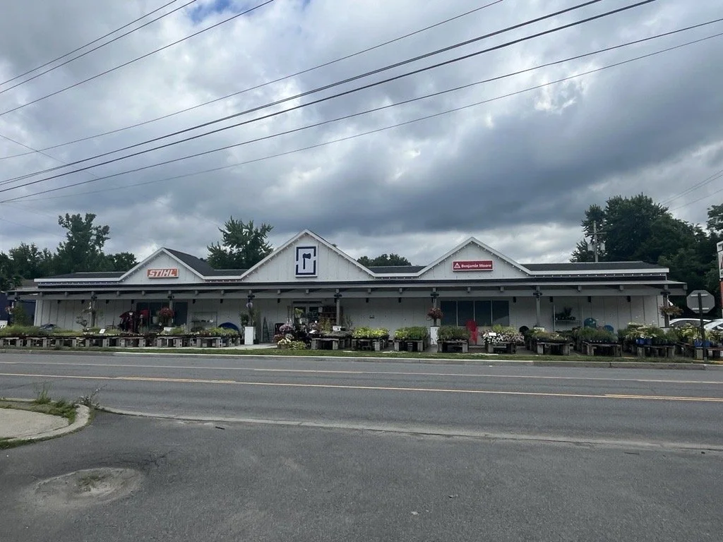 A single-story building with a white exterior, displaying signs for STIHL, Benjamin Moore, and another brand, with outdoor flower displays and wheels lined up in front of the building under a cloudy sky.