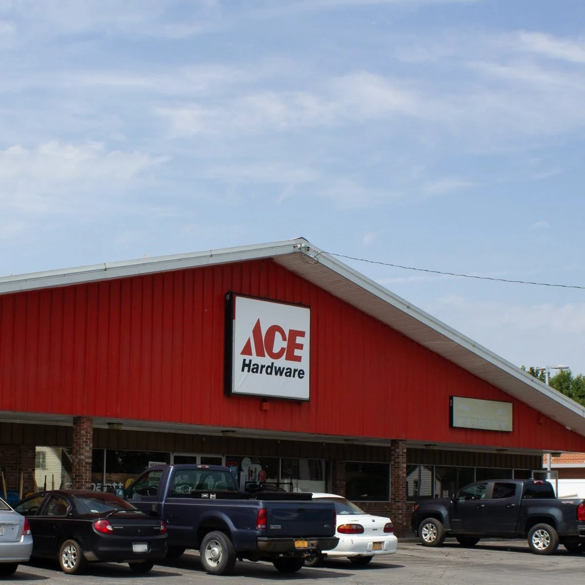 Exterior of an ACE Hardware store with a red building, parked cars in front, and a blue sky with clouds above.