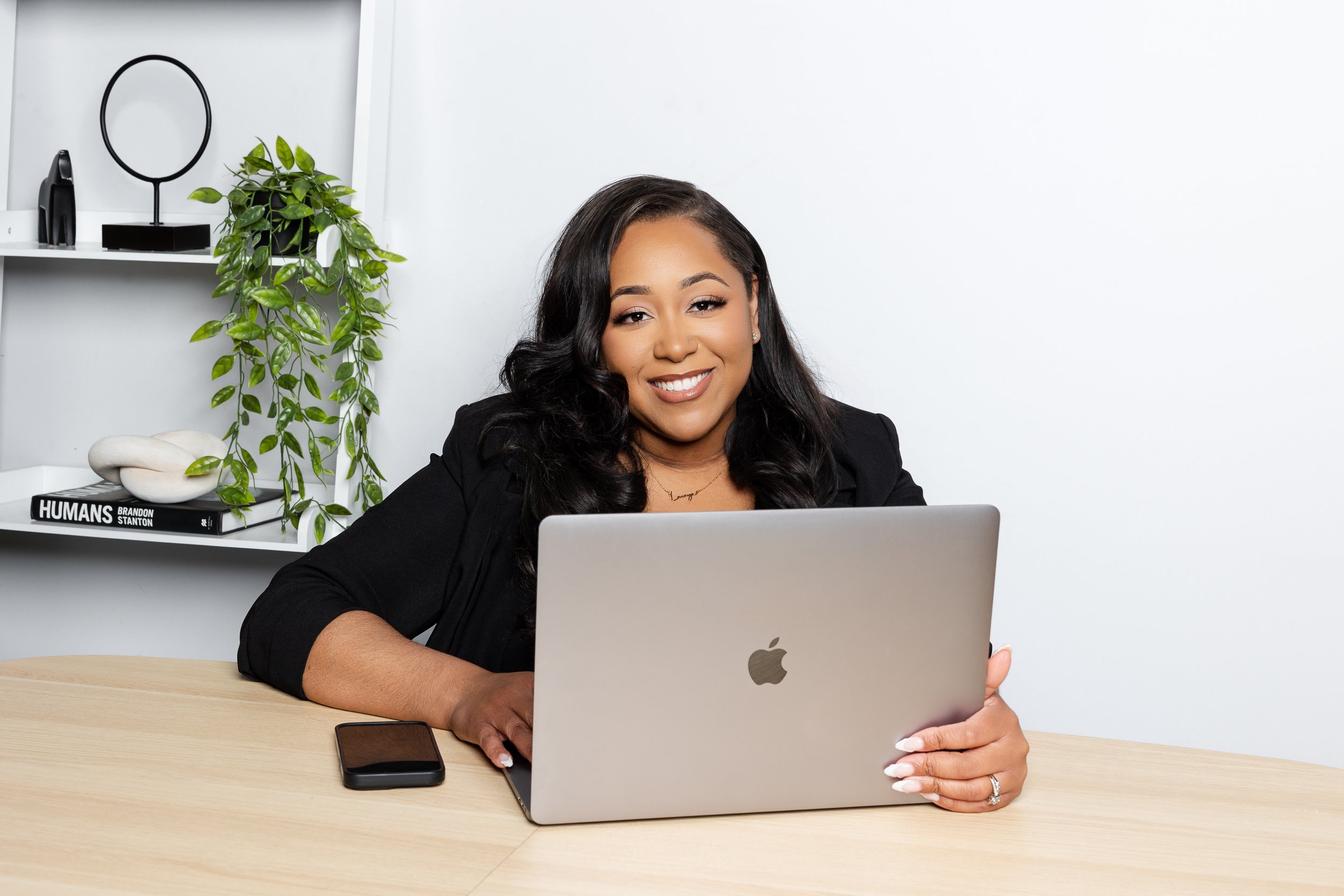A woman with dark hair and a black blazer sitting at a light wooden desk, smiling and looking at a silver laptop. A black smartphone and a stylus are on the desk. Behind her, a white shelf holds a green potted plant, a black circular mirror, and a black decorative object. A book titled 'HUMANS' by Brandon Stanton is also on the shelf.